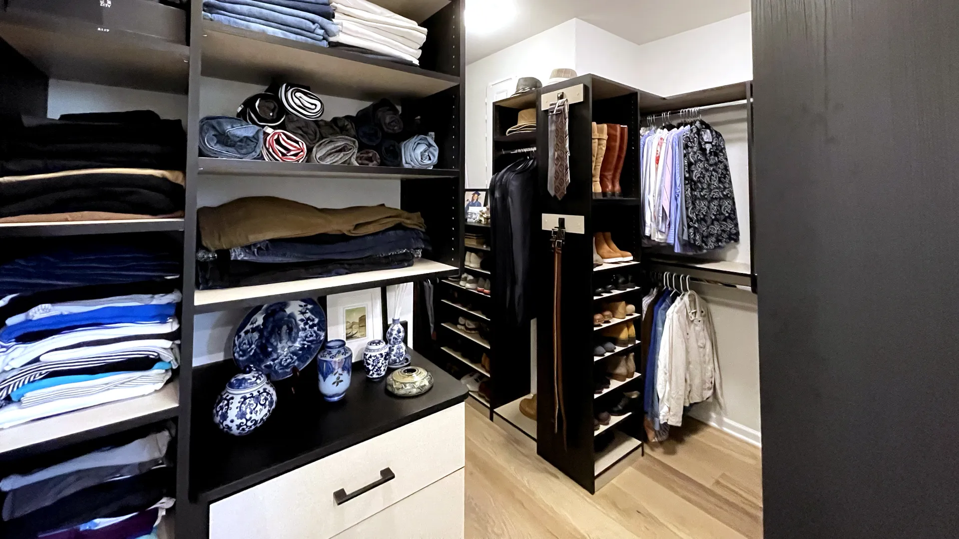 Organized walk-in closet with shelves of folded clothes, hanging garments, boots, and decorative blue-and-white porcelain items.