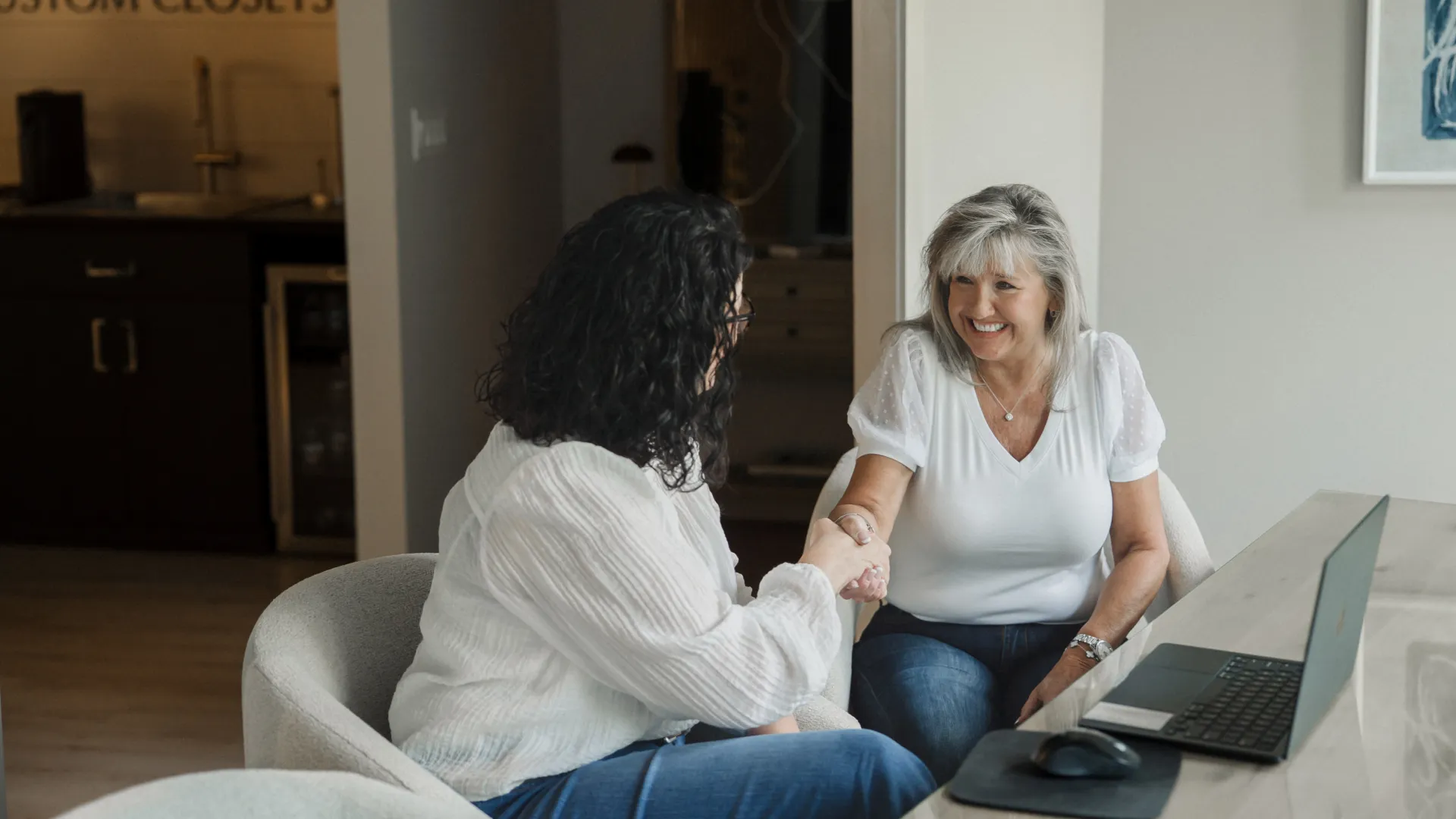 Two women shaking hands in a modern office setting with a laptop and smiling during a meeting.