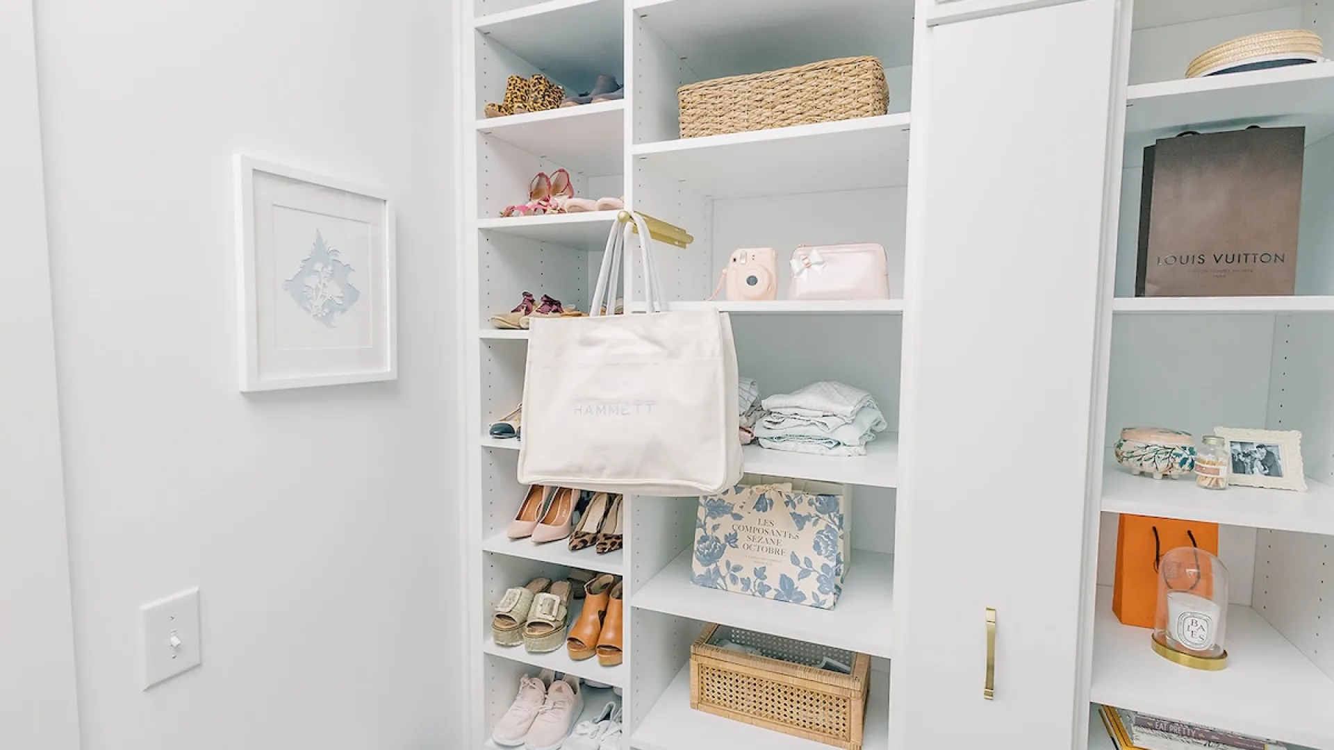 Organized white shelving with shoes, a hanging tote bag, wicker baskets, folded clothes, and decorative items in a closet