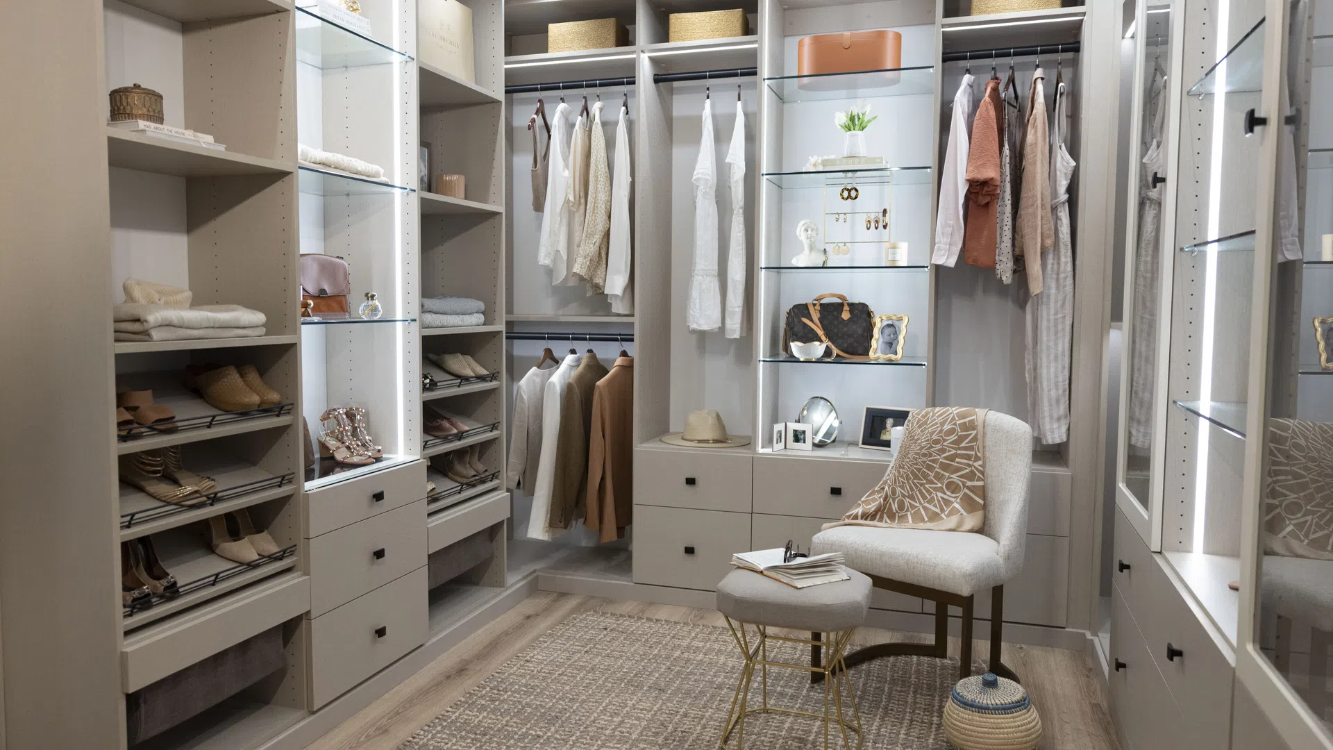 Modern walk-in closet with organized shelves, hanging clothes, shoe racks, a chair, and neutral decor accents