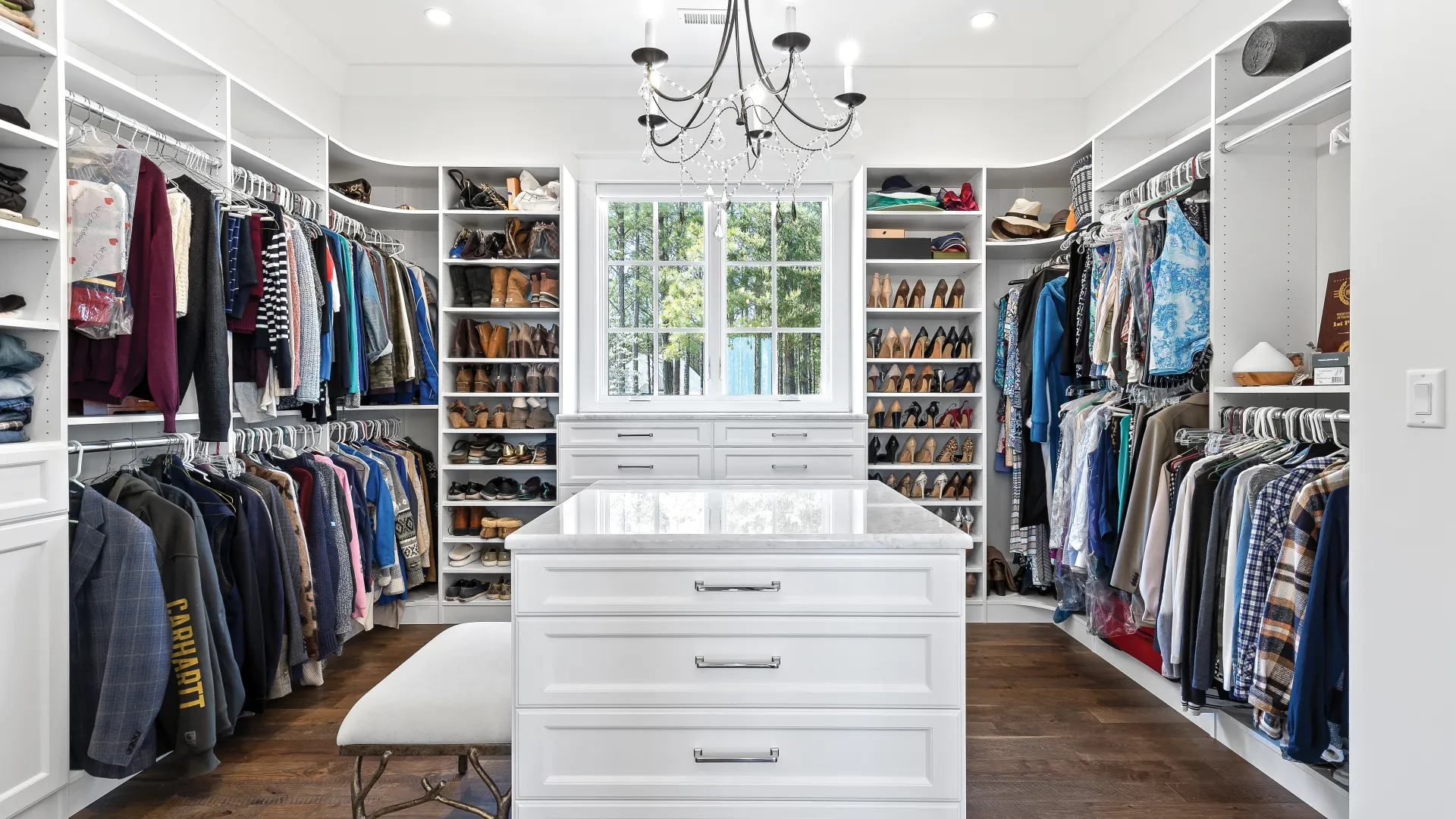 Spacious white walk-in closet with a marble island, hanging clothes, shoe shelves, chandelier, and window with outdoor view.