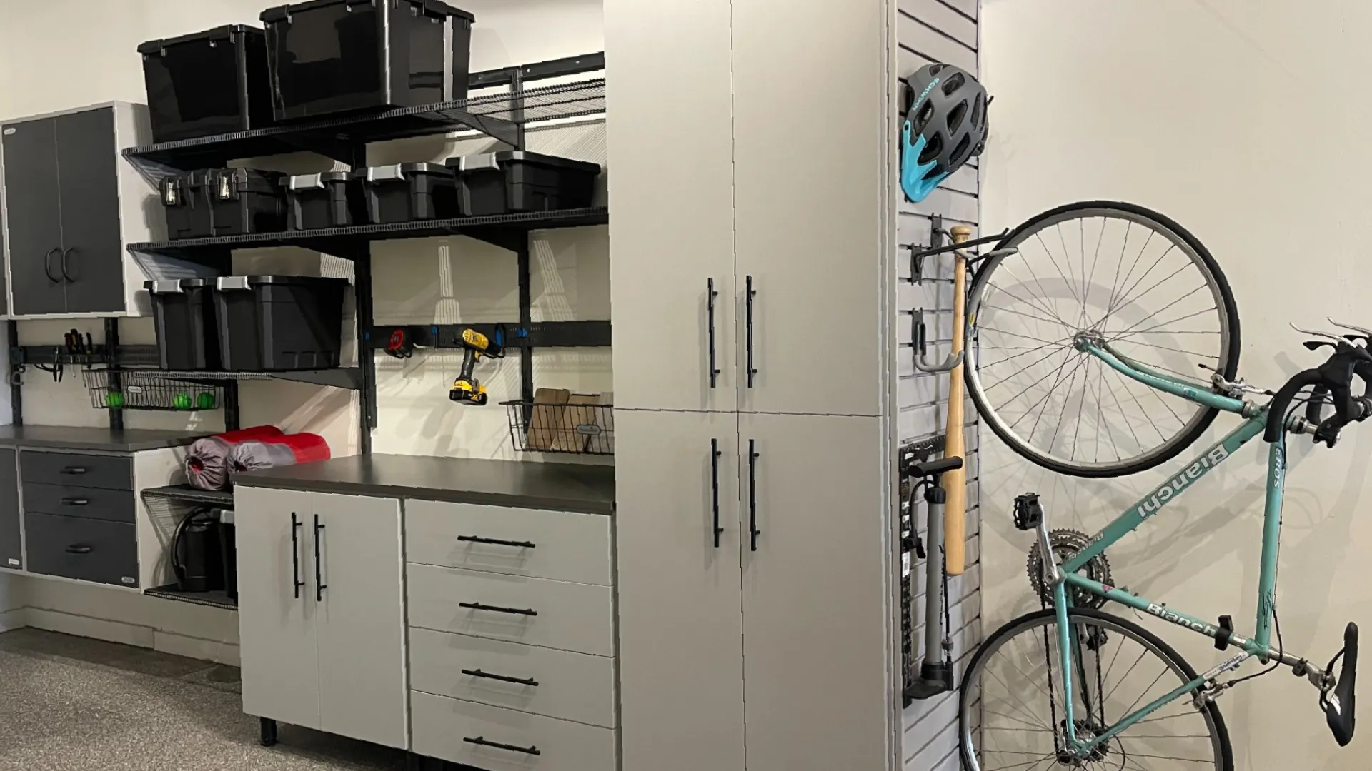 Organized garage with gray storage cabinets, black bins on shelves, and a vertical bike rack holding a teal bicycle and helmet.