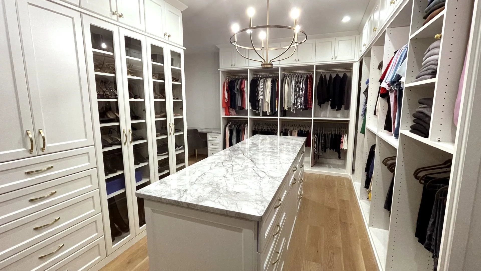 Spacious walk-in closet with marble island, white cabinets, glass doors, shoes, hanging clothes, light fixture and hardwood floors