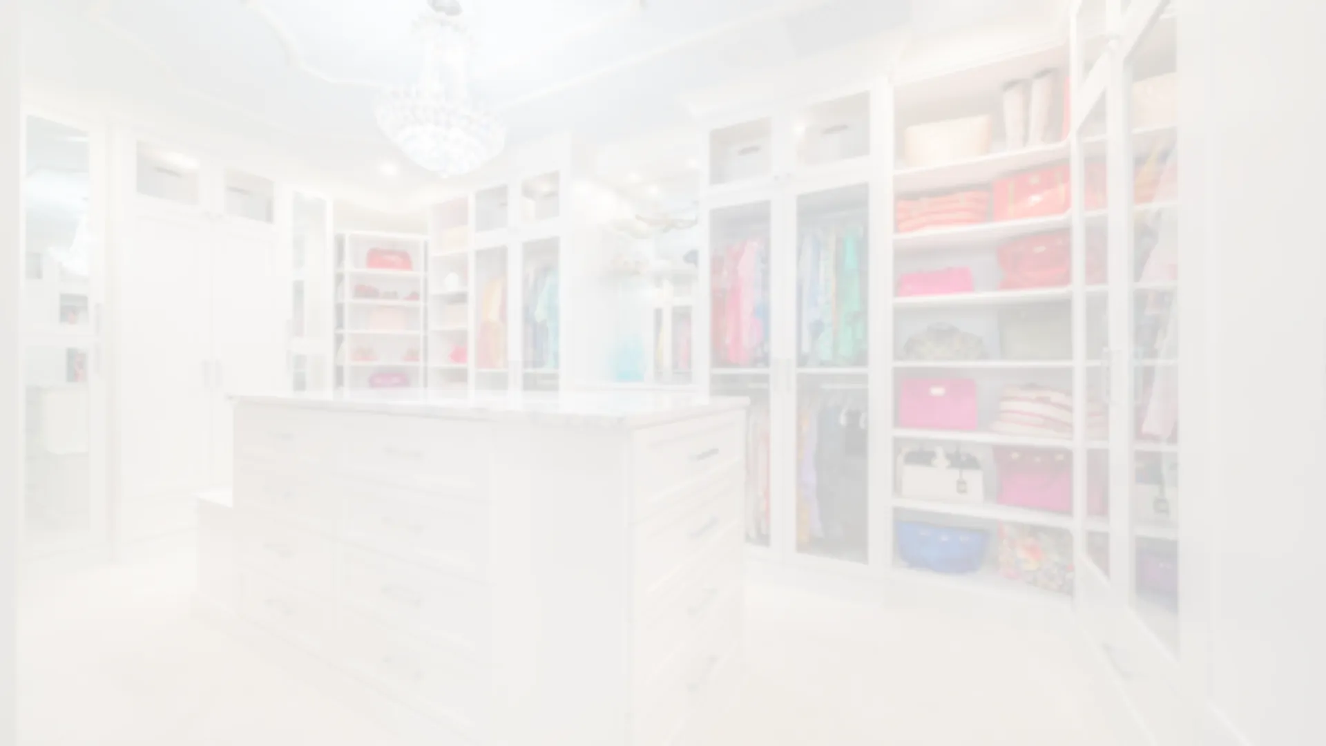 Spacious white walk-in closet with glass cabinets, organized shelves, island drawers, and a chandelier light fixture