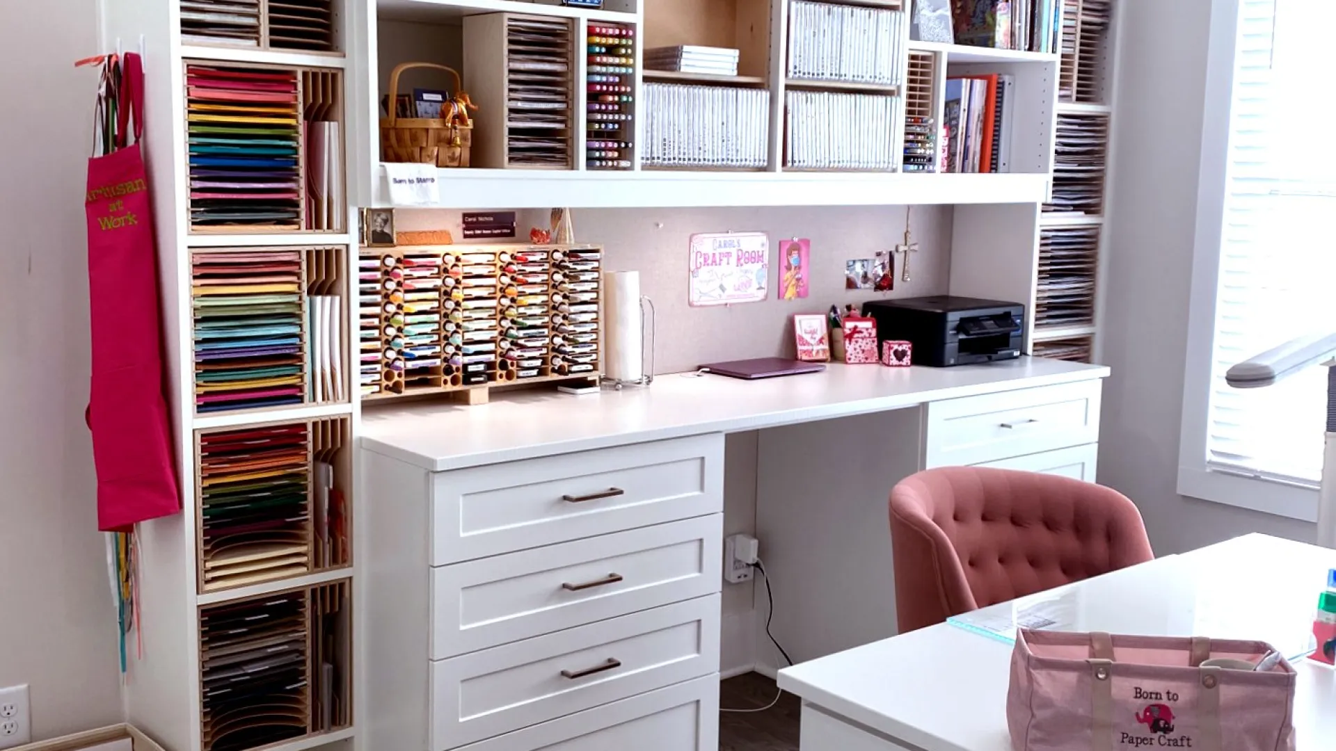 Organized craft room with white cabinets, white desk, drawers, scrapbook supplies, crafting supplies and a pink cushioned chair, hardwood flooring