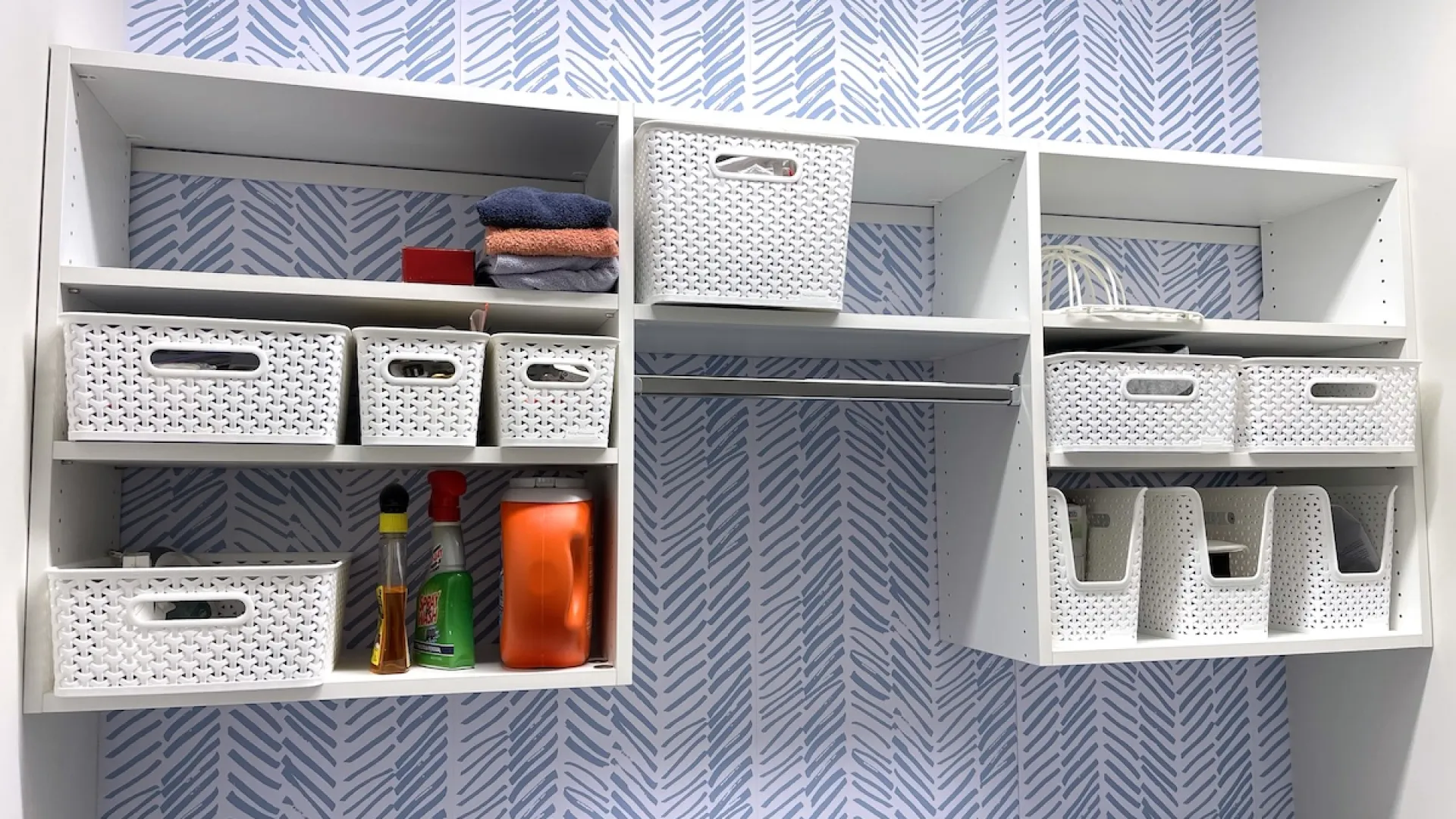White wall-mounted shelving with woven storage baskets and cleaning supplies against blue patterned wallpaper