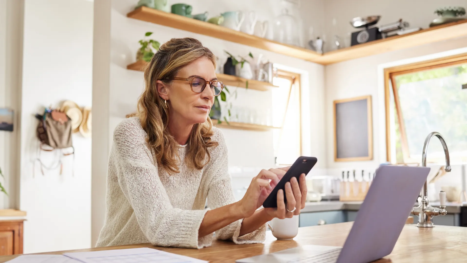 Woman in glasses using smartphone and laptop at kitchen table with papers and coffee mug