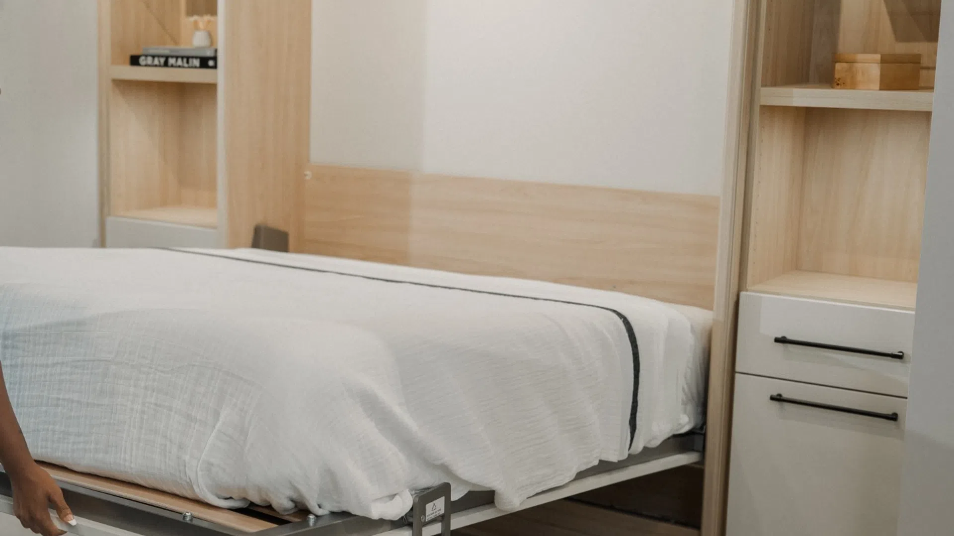 Woman pulling down a Murphy bed in a modern bedroom with light wood shelves and stacked washer dryer.