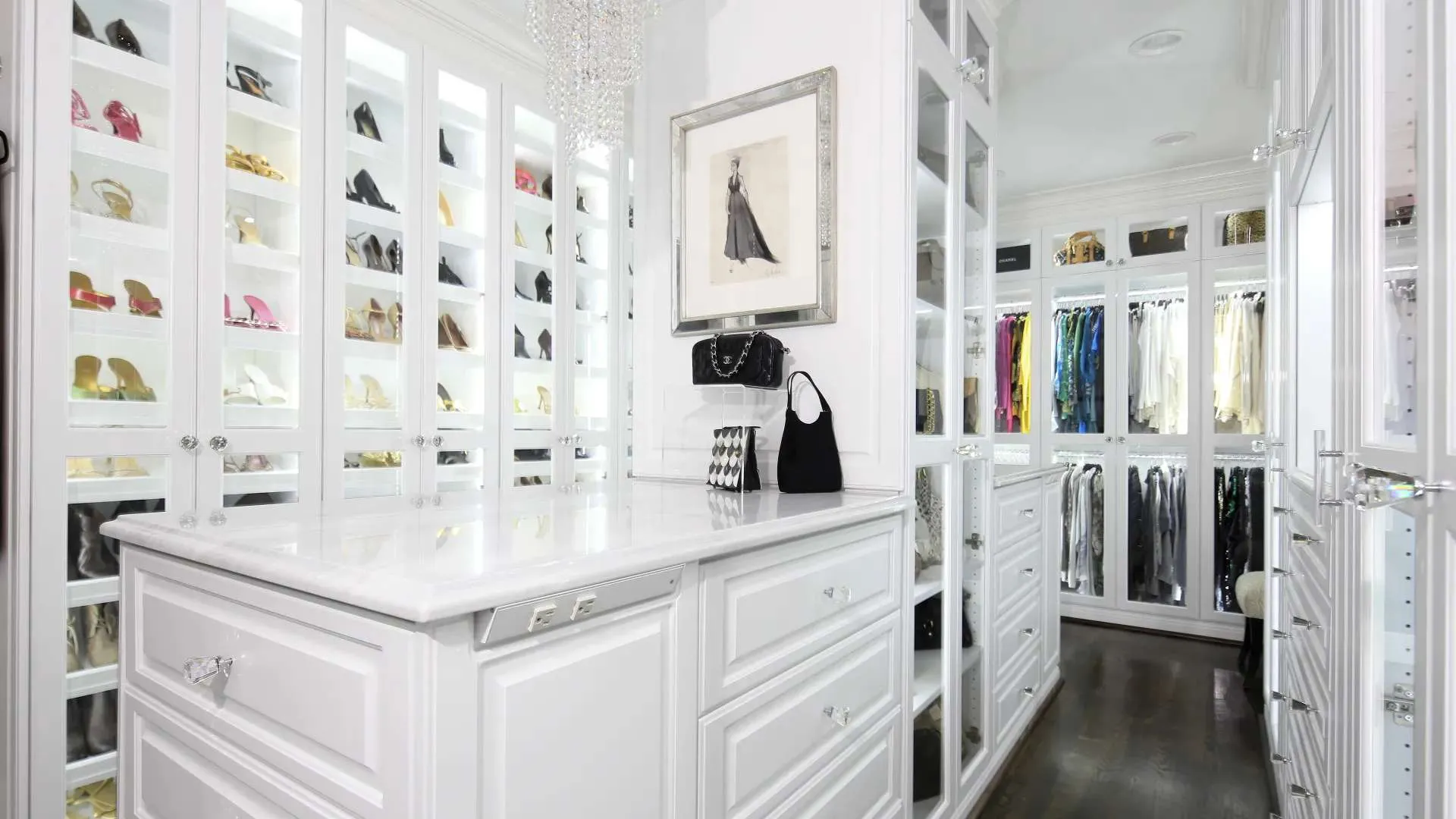 Bright white walk-in closet with glass cabinets displaying shoes and hanging clothes with marble island counter.