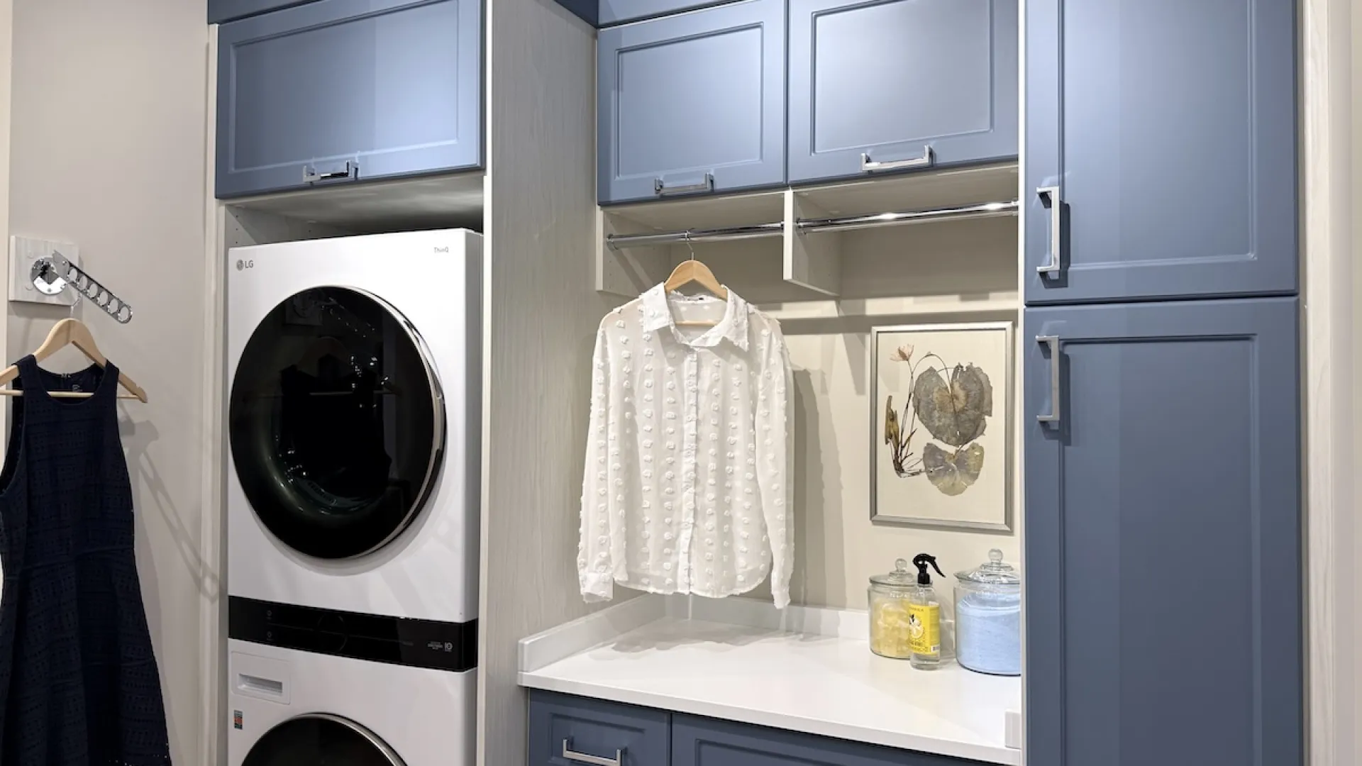 Modern laundry room with stacked washer dryer, blue cabinets, hanging shirt, and countertop with storage jars.