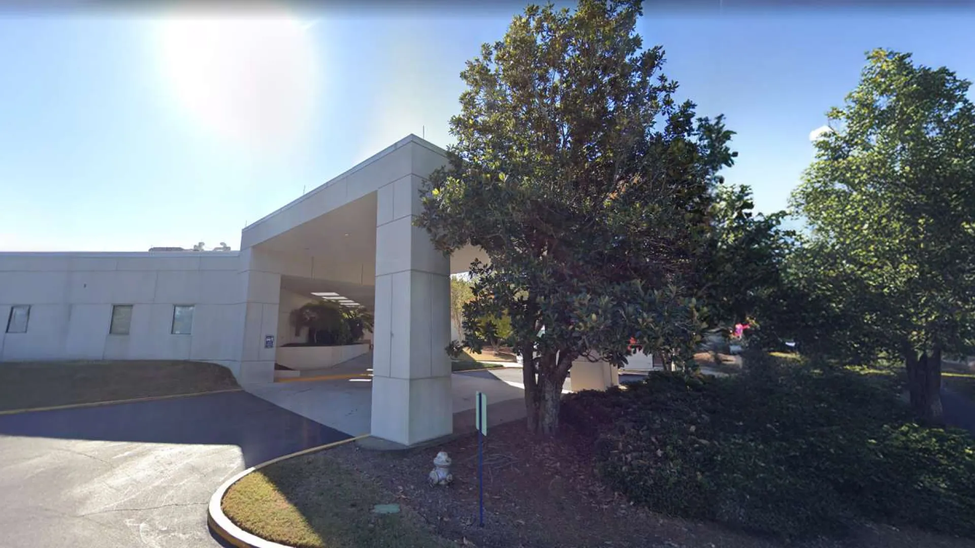Modern white building with a covered entrance, surrounded by green trees and a curved driveway in daylight.