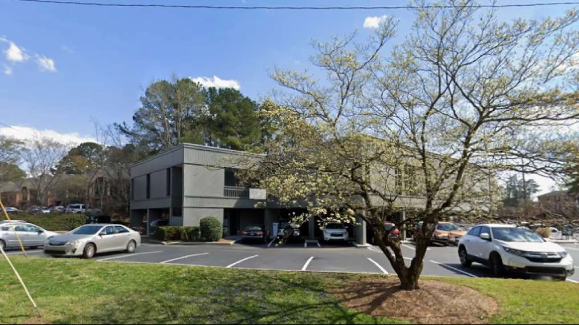 Modern two-story office building with parking lot, cars, and a large tree in a sunny suburban setting.