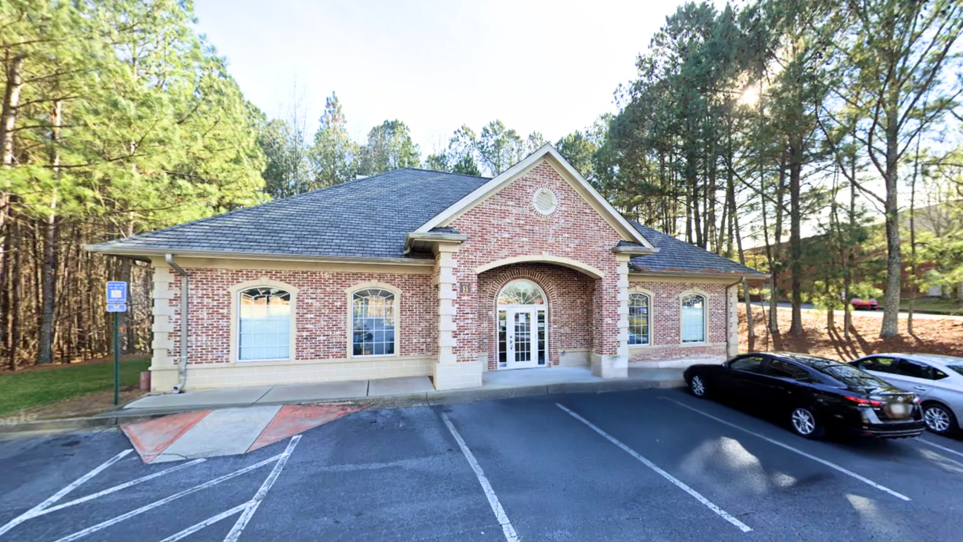 Brick office building with arched entrance surrounded by trees and parked cars in front under a clear sky