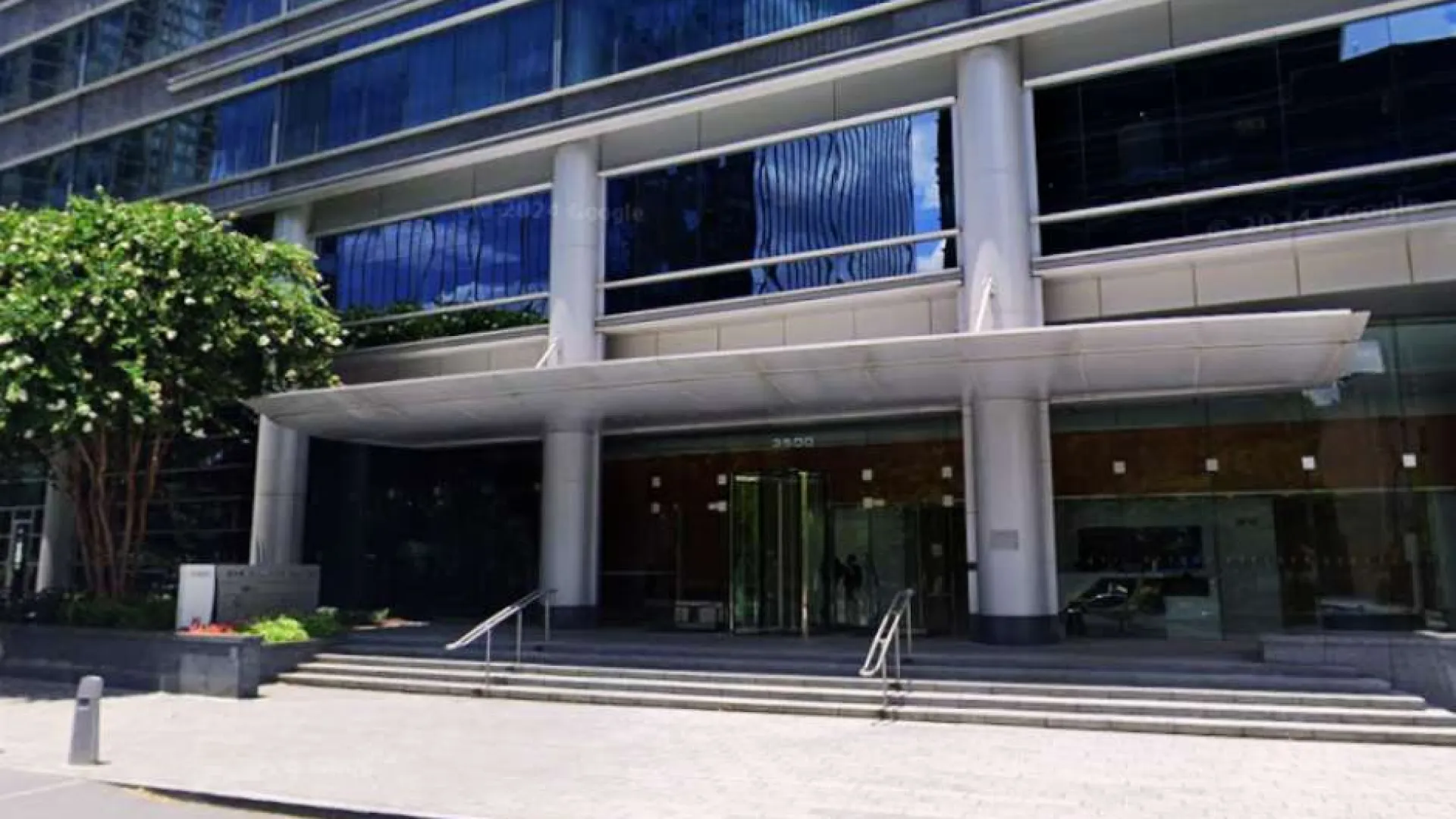 Modern office building entrance with glass facade, steps, and large white pillars surrounded by trees and pavement.