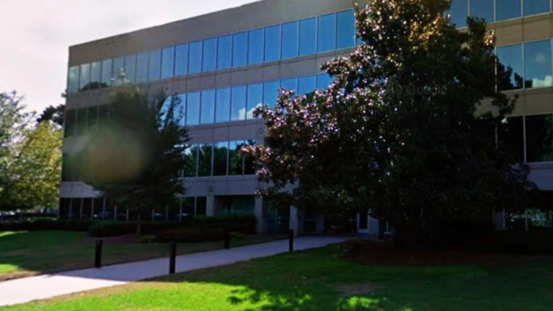 Modern glass office building with reflective windows, surrounded by trees and a well-maintained lawn on a sunny day