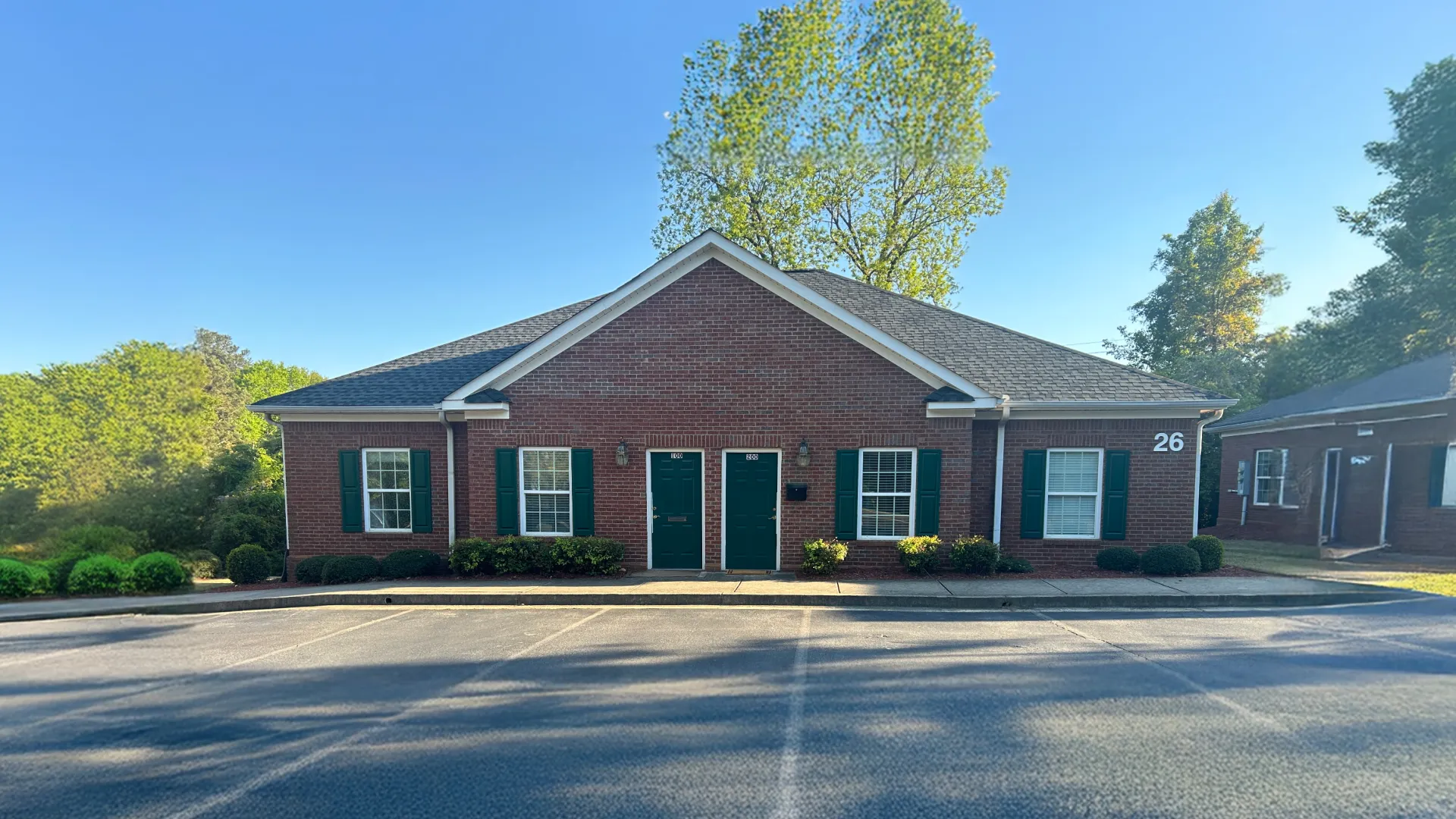 Single-story red brick duplex with green doors and shutters surrounded by trees and bushes under clear sky