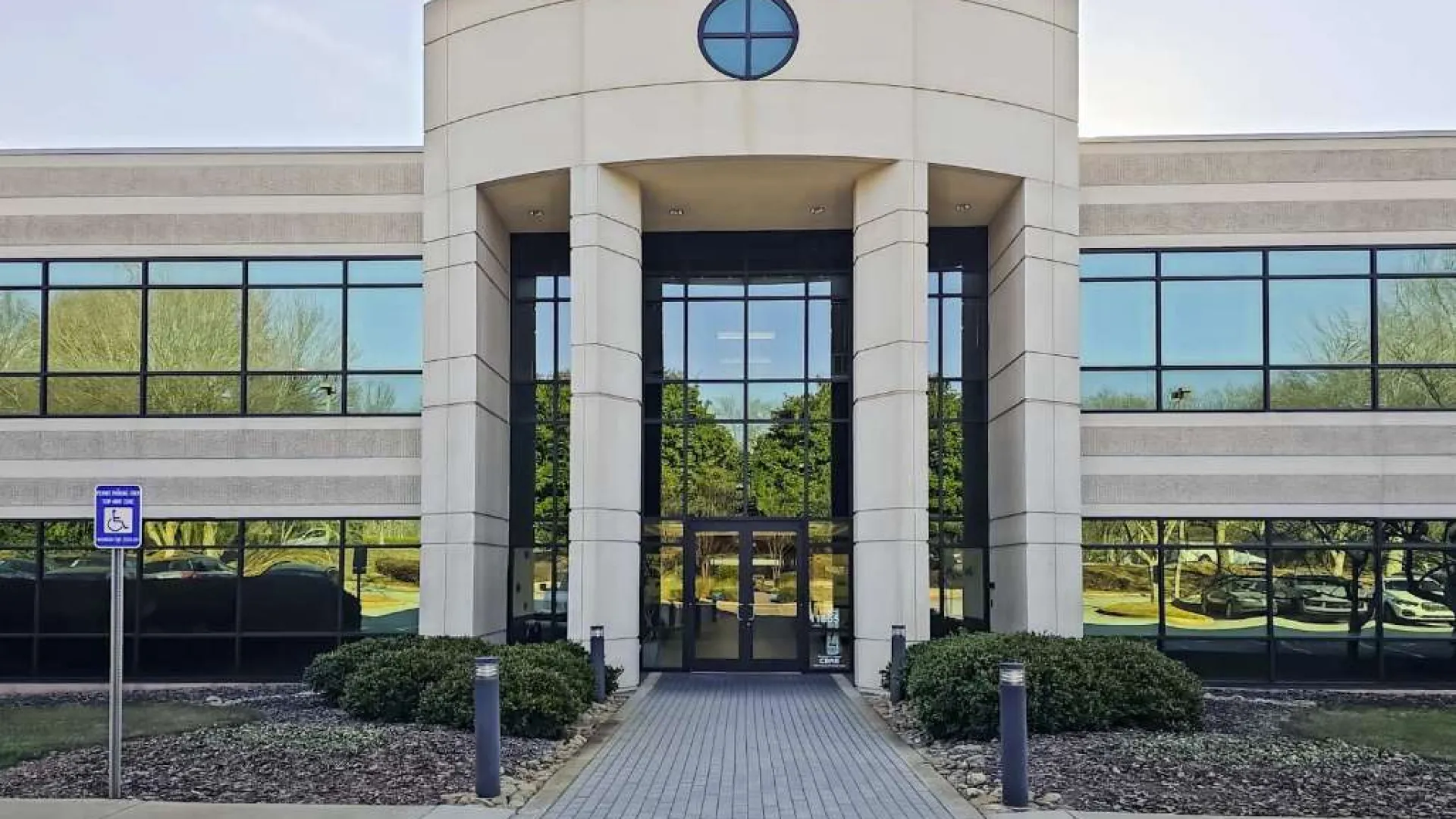 Modern office building entrance with large glass windows, stone pillars, and landscaped greenery in front.