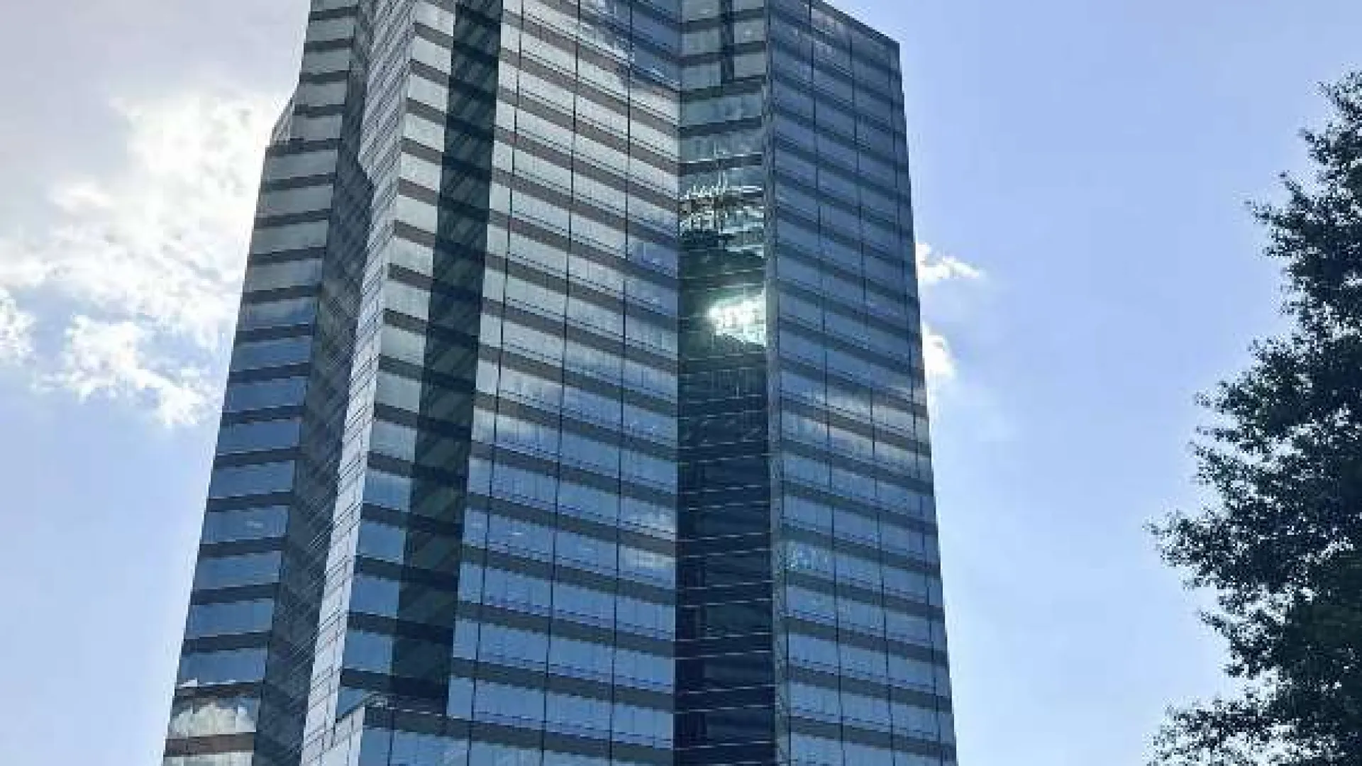 Modern glass skyscraper reflecting sky and clouds with trees in the foreground under a clear blue sky.