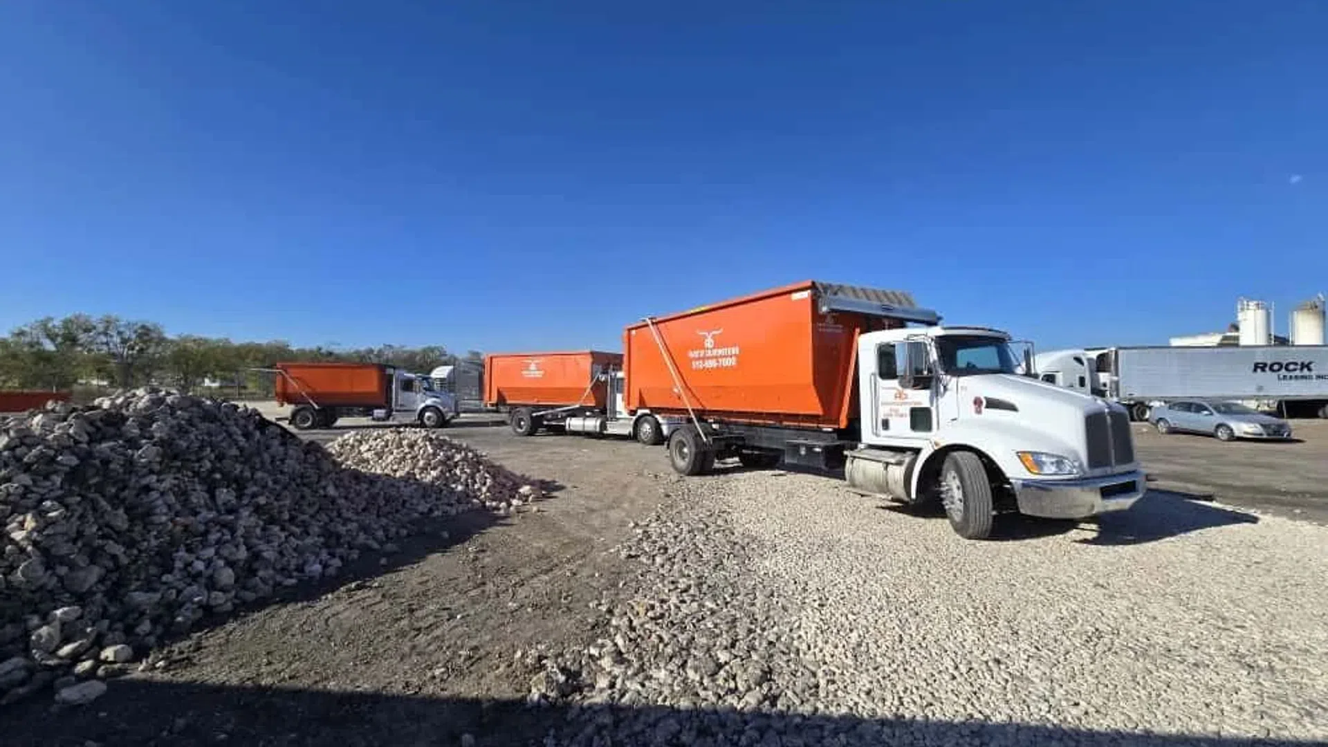 White trucks with bright orange containers parked on gravel near stone piles under clear blue sky.