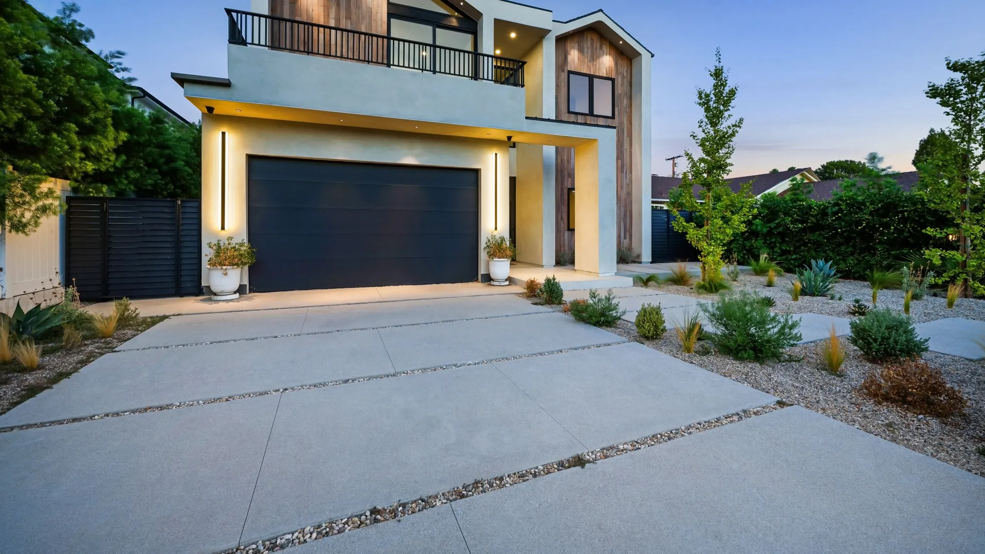 Modern two-story house with sleek lines, dark garage door, and landscaped garden at dusk.