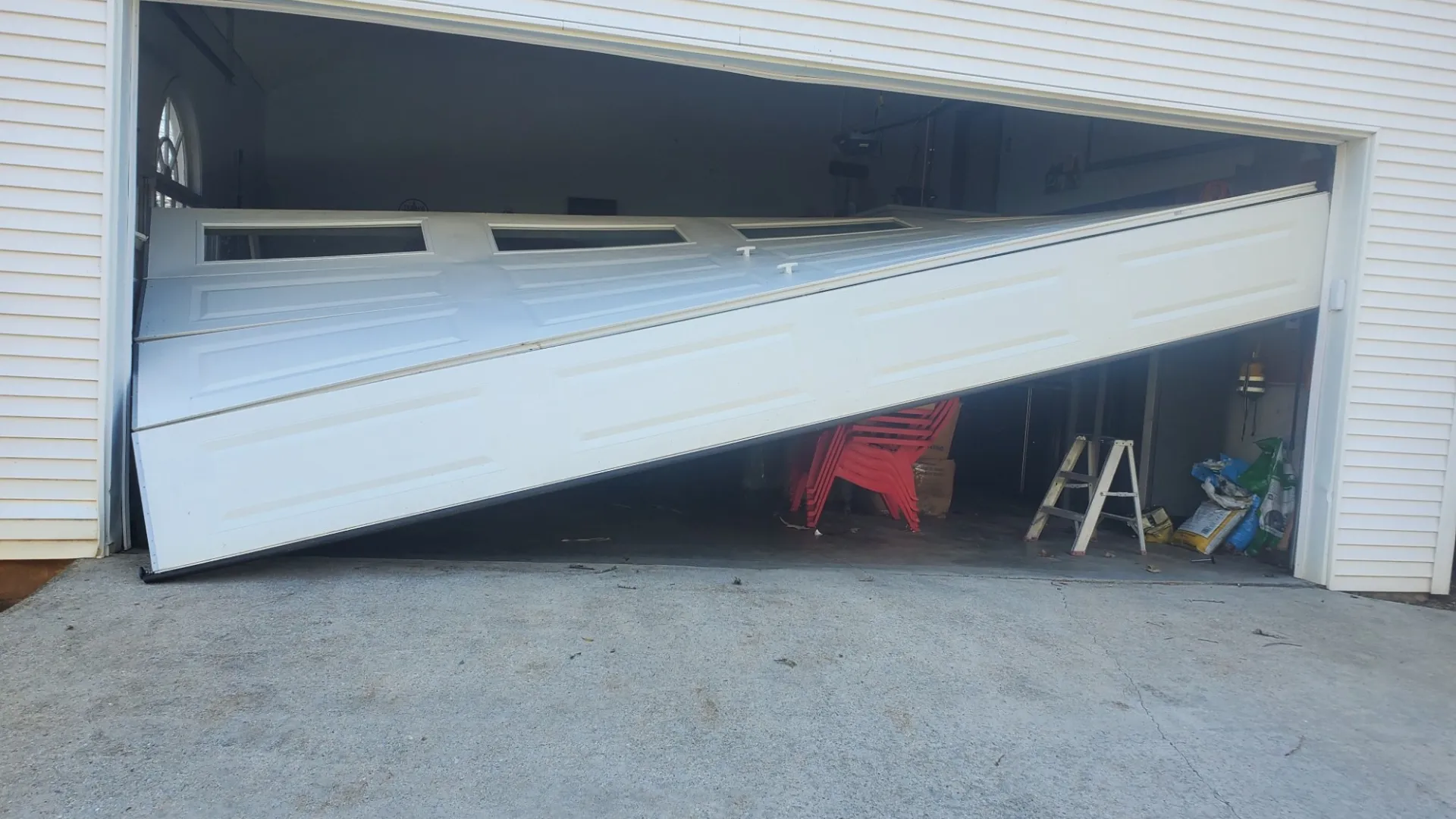 A damaged garage door partially collapsed into the garage, revealing tools and a ladder inside.