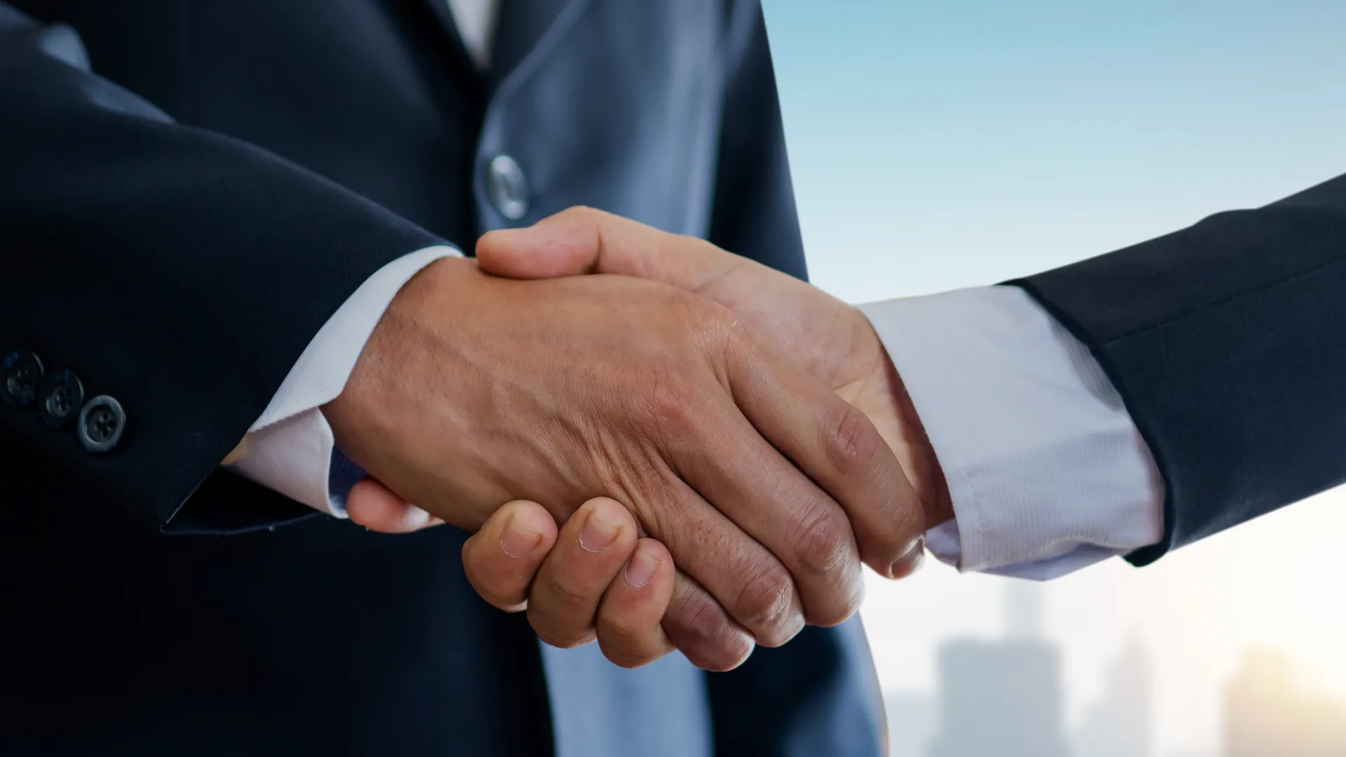 Two business professionals shaking hands in formal suits with city skyline in the background at sunrise.