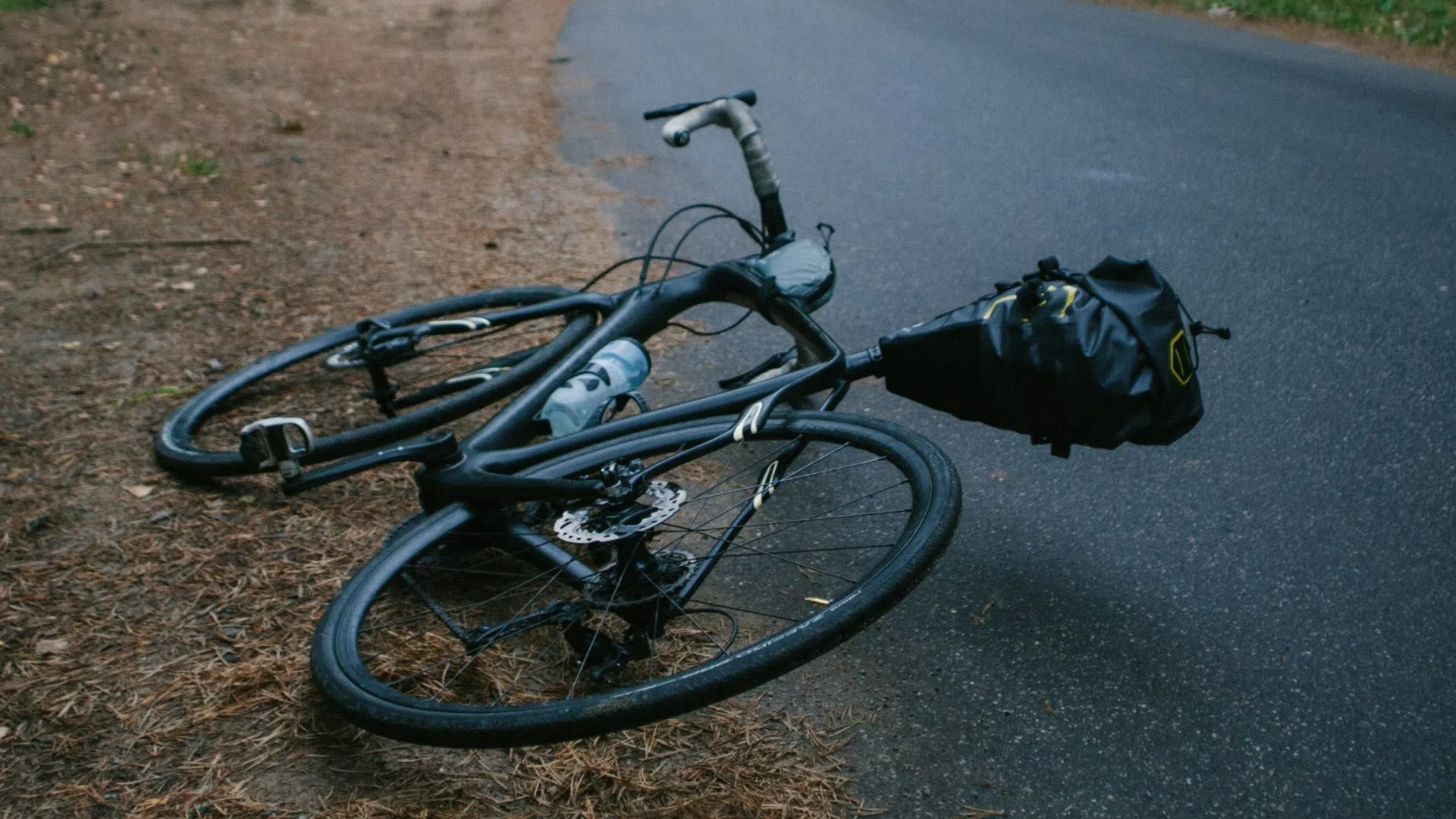 Road bike with pannier bag lying on the forest road edge surrounded by trees and dirt.