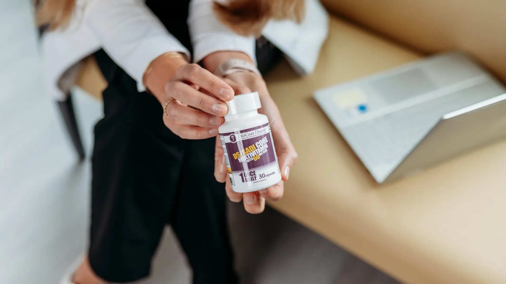 Person holding a bottle of dietary supplement with a laptop in the background on a couch.