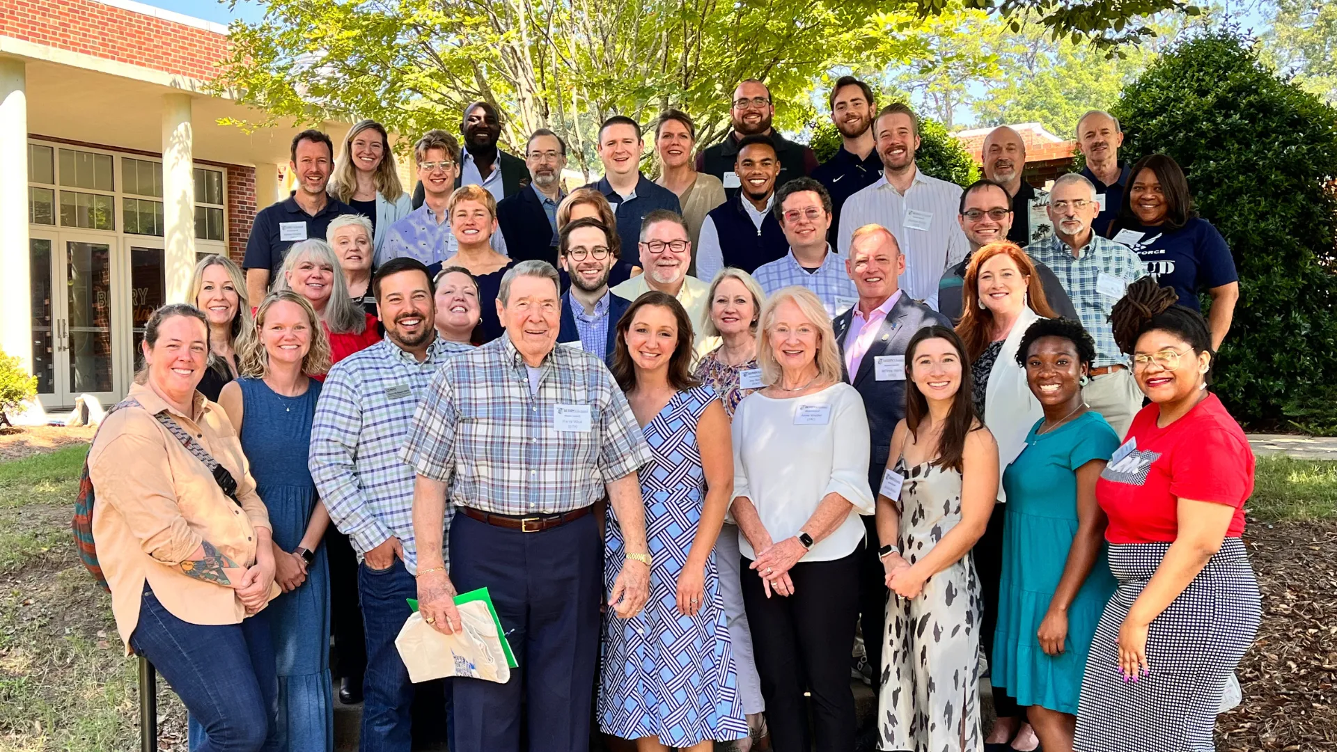 Large diverse group of people smiling outdoors in front of building and trees on a sunny day