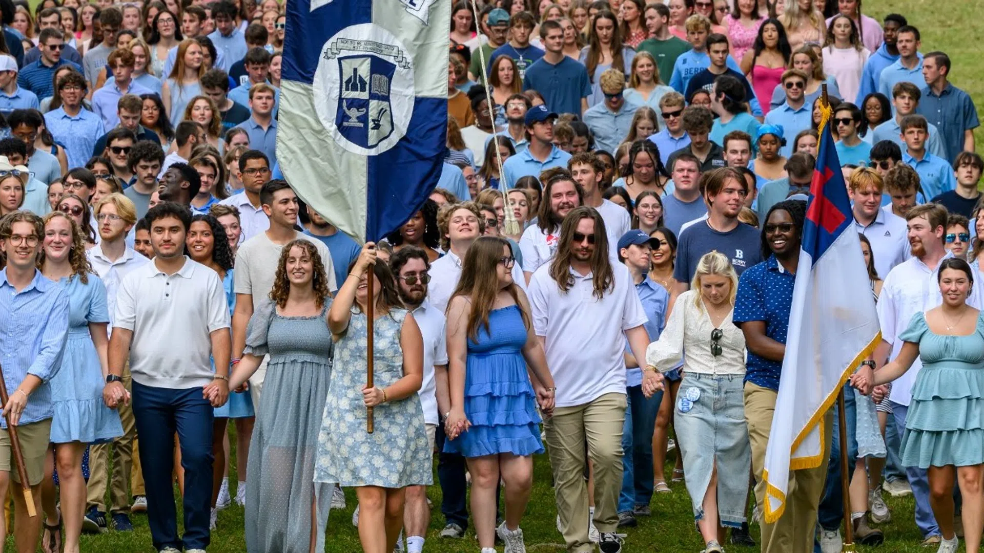 Large group of Berry College students walking outdoors holding flags during an organized event on green lawn.