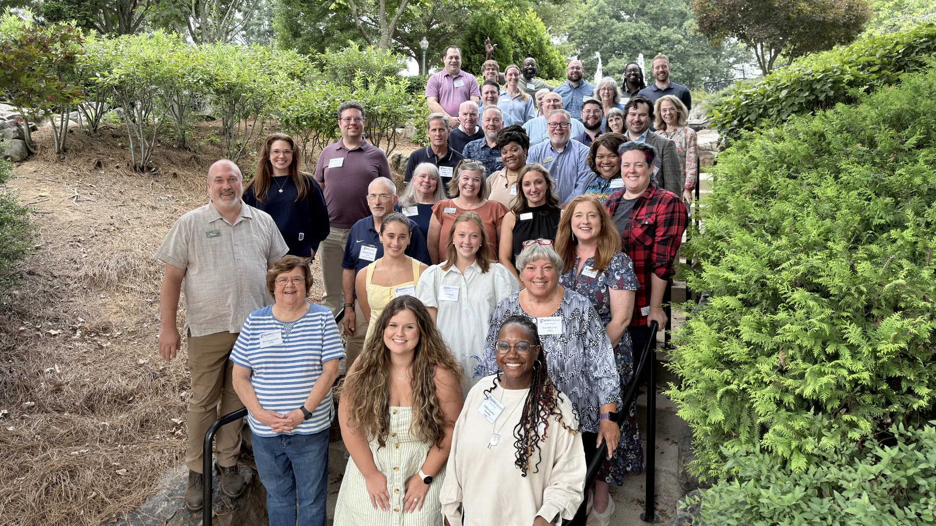 Group photo of diverse adults smiling on outdoor stairs surrounded by trees and greenery on a sunny day