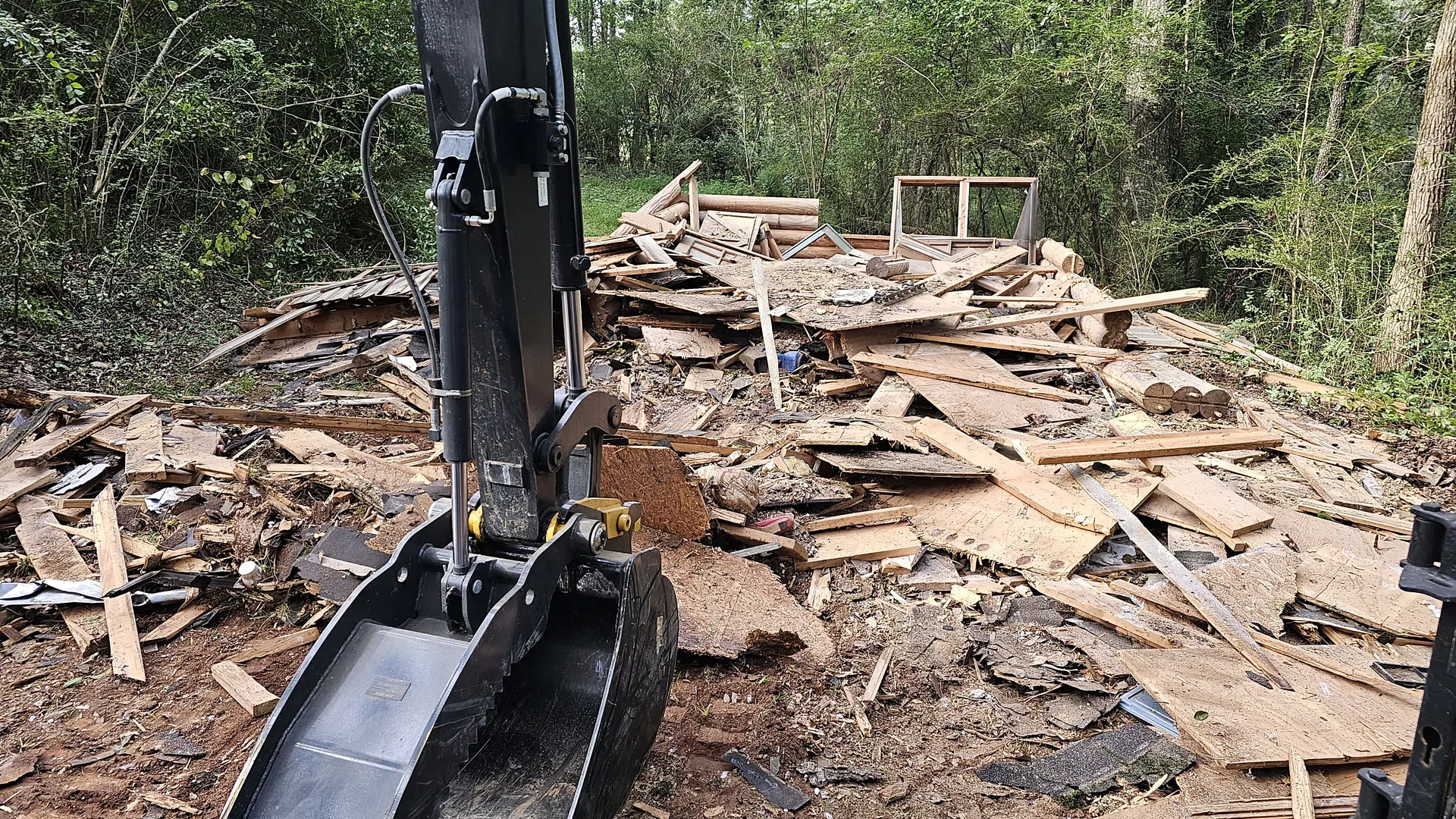 Excavator bucket next to a pile of broken wooden debris in a forest clearing under construction.