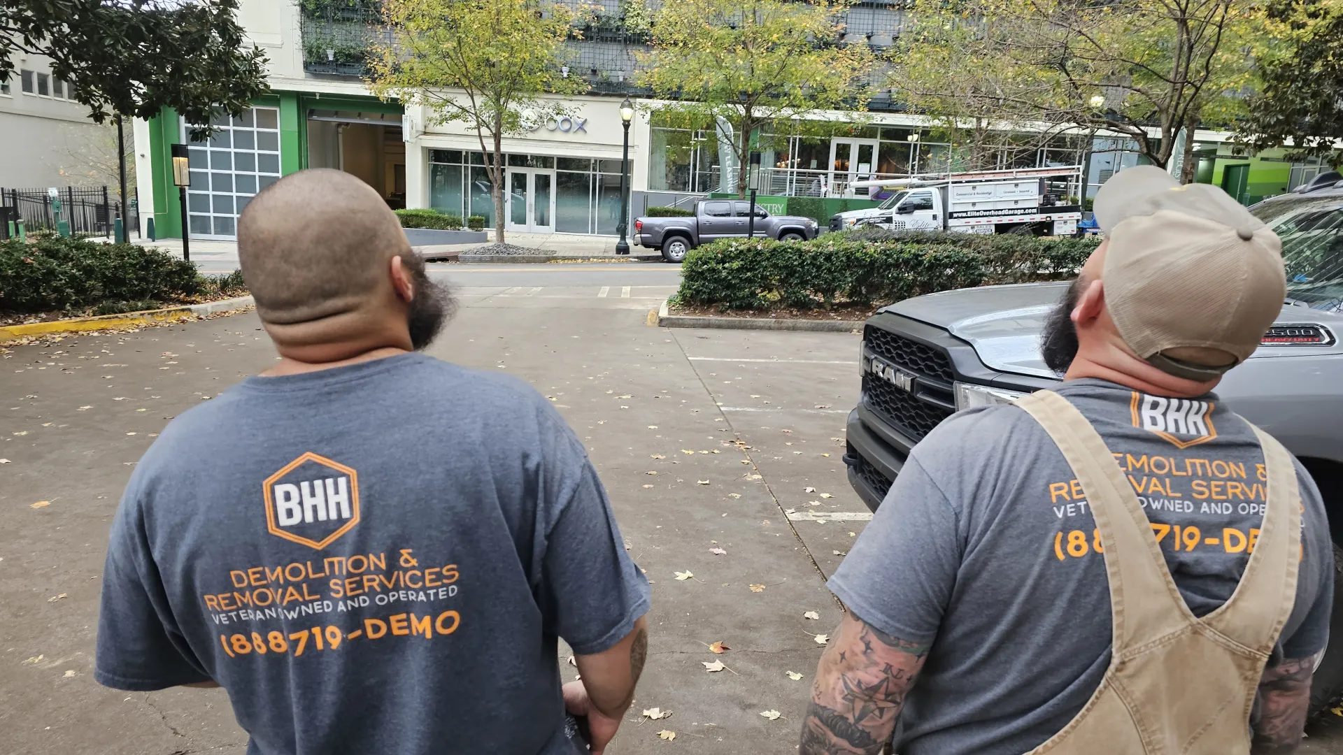 Two workers wearing BHH Demolition & Removal Services t-shirts standing near trucks in an urban outdoor setting