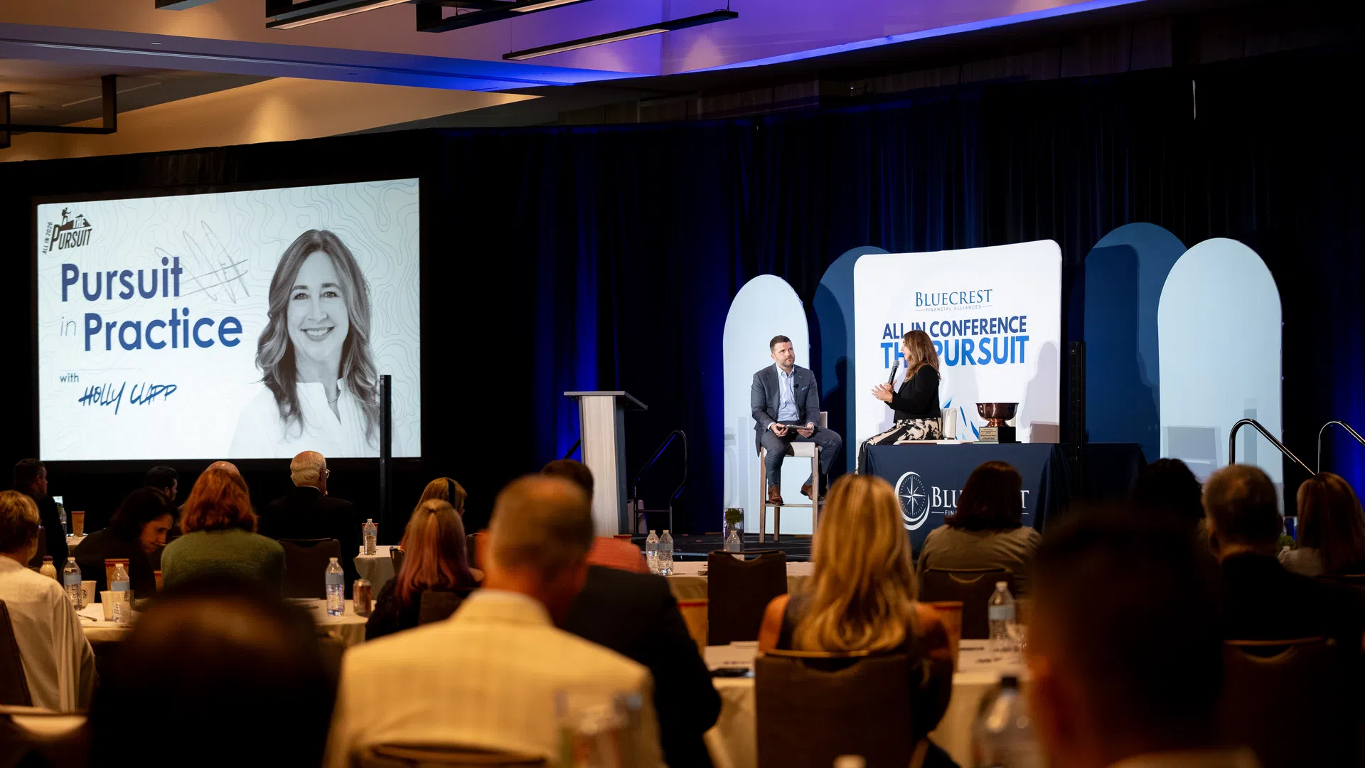 Conference attendees watch a live interview on stage with a large screen displaying Pursuit in Practice branding.