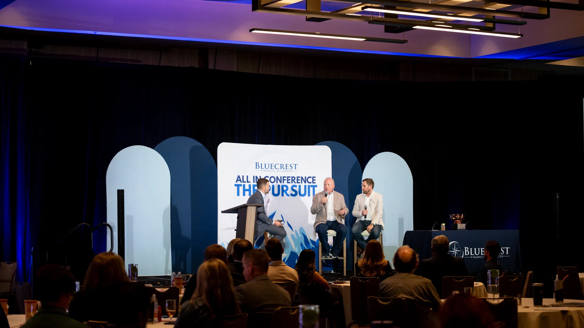 Three men on stage speaking at the Bluecrest All In Conference with an audience watching in a dimly lit room