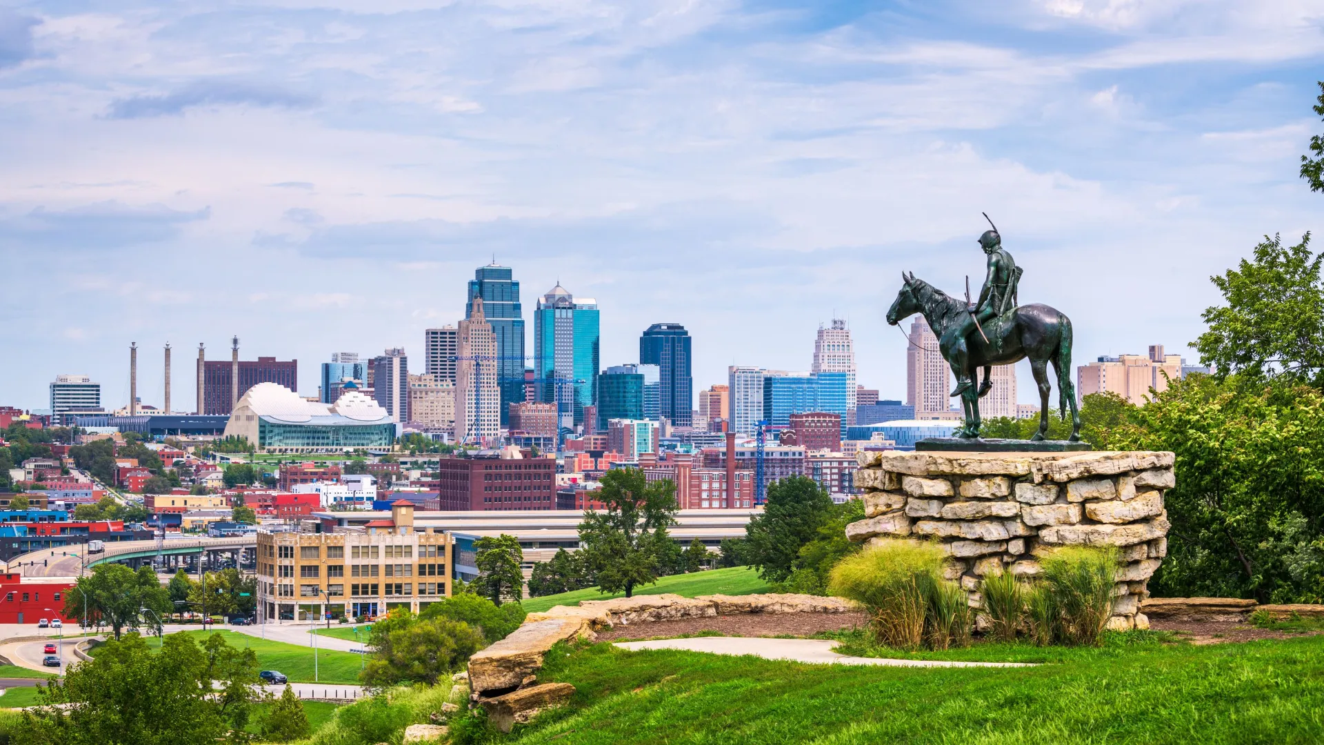 a statue of a person riding a horse in a park with a city in the background