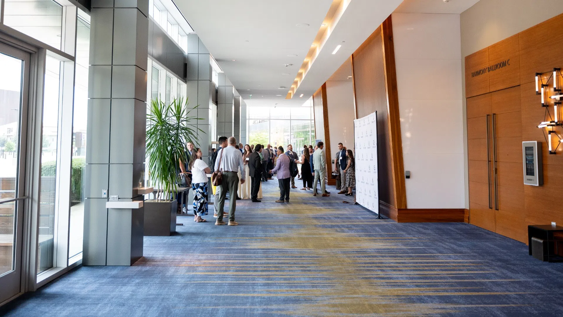 Modern conference hallway with groups of people networking near large windows and wooden wall panels.