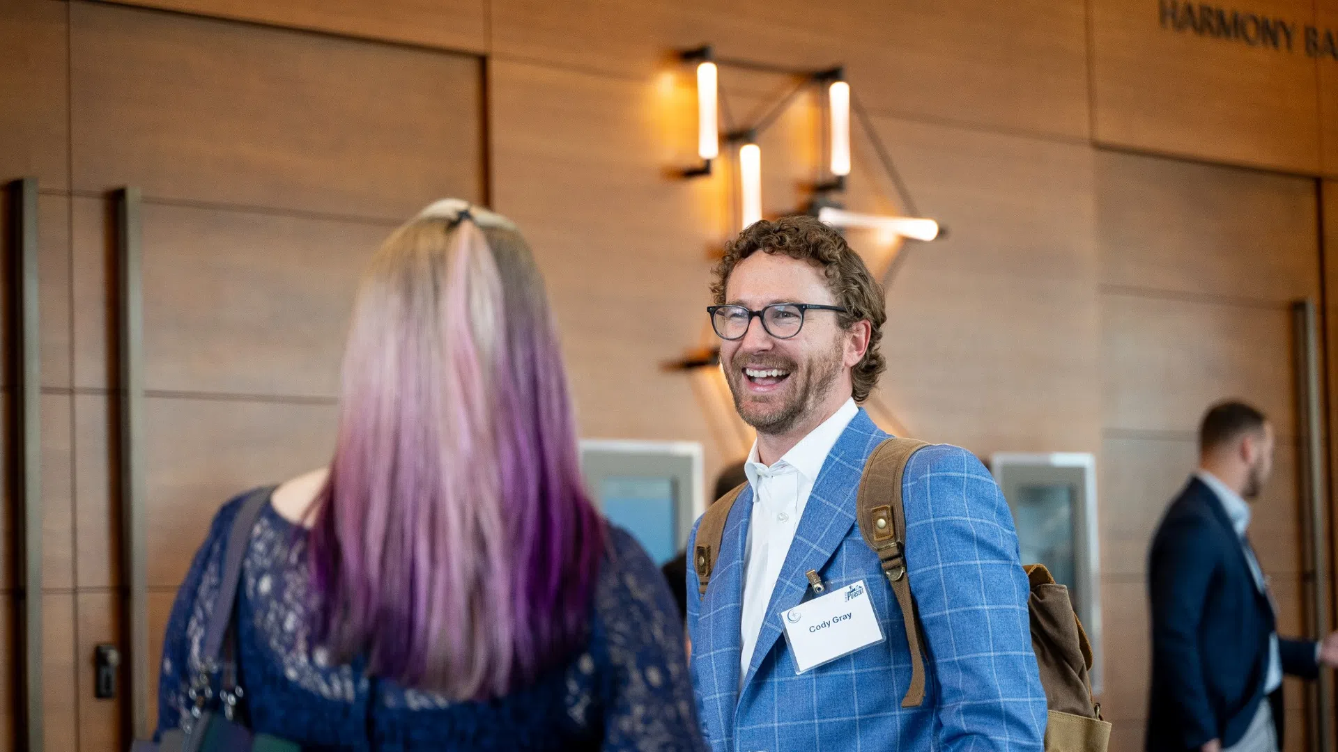 Man in blue checkered blazer smiling and talking with woman with purple hair in a modern conference lobby.