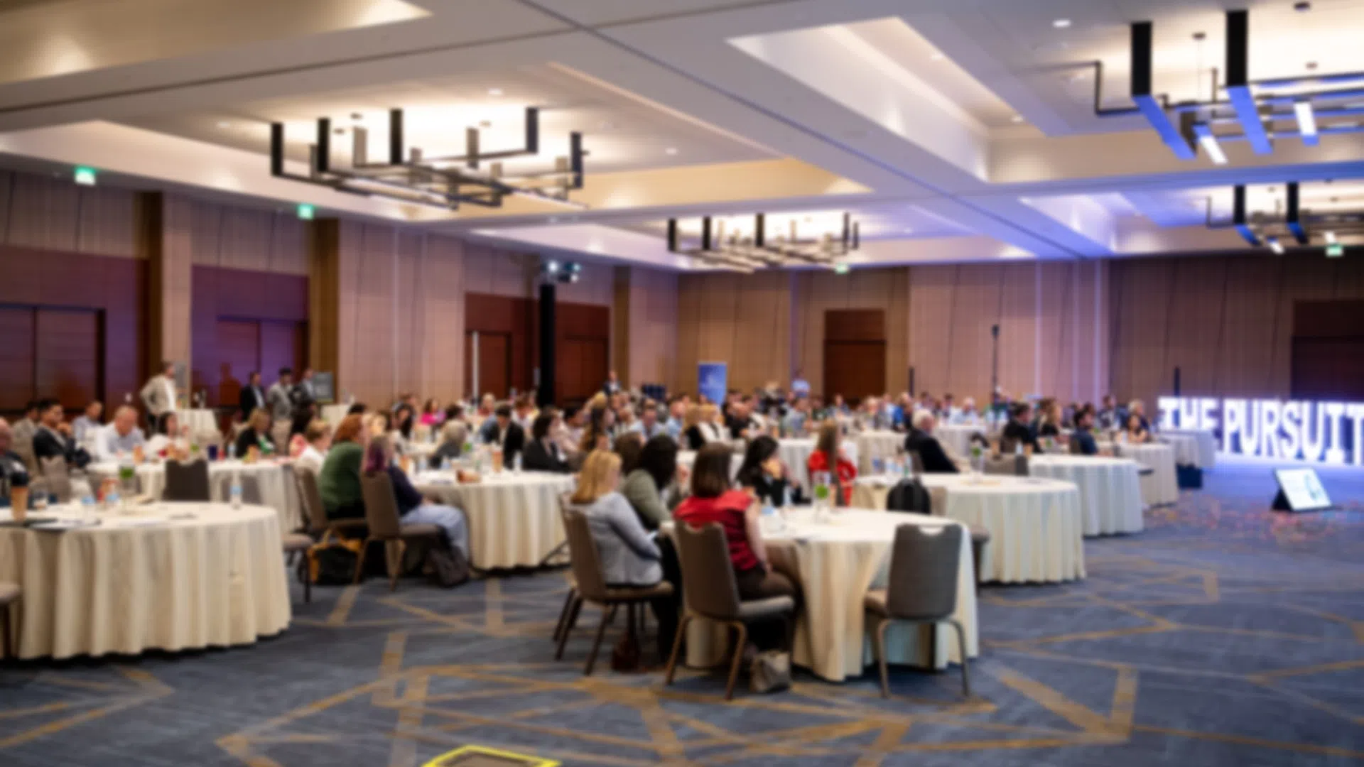 Large conference room with attendees seated at round tables listening to a presentation with THE PURSUIT sign.