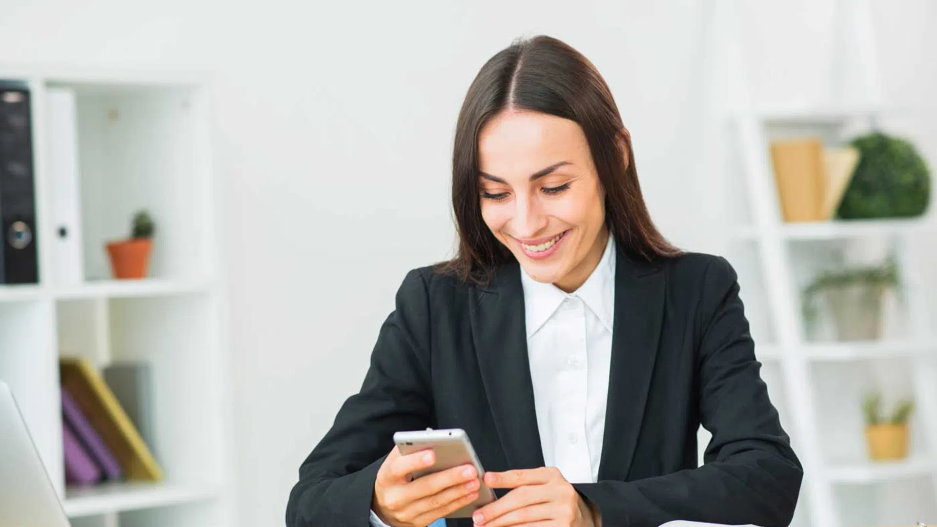 Smiling businesswoman in black suit using smartphone at desk with laptop and open notebook in office.