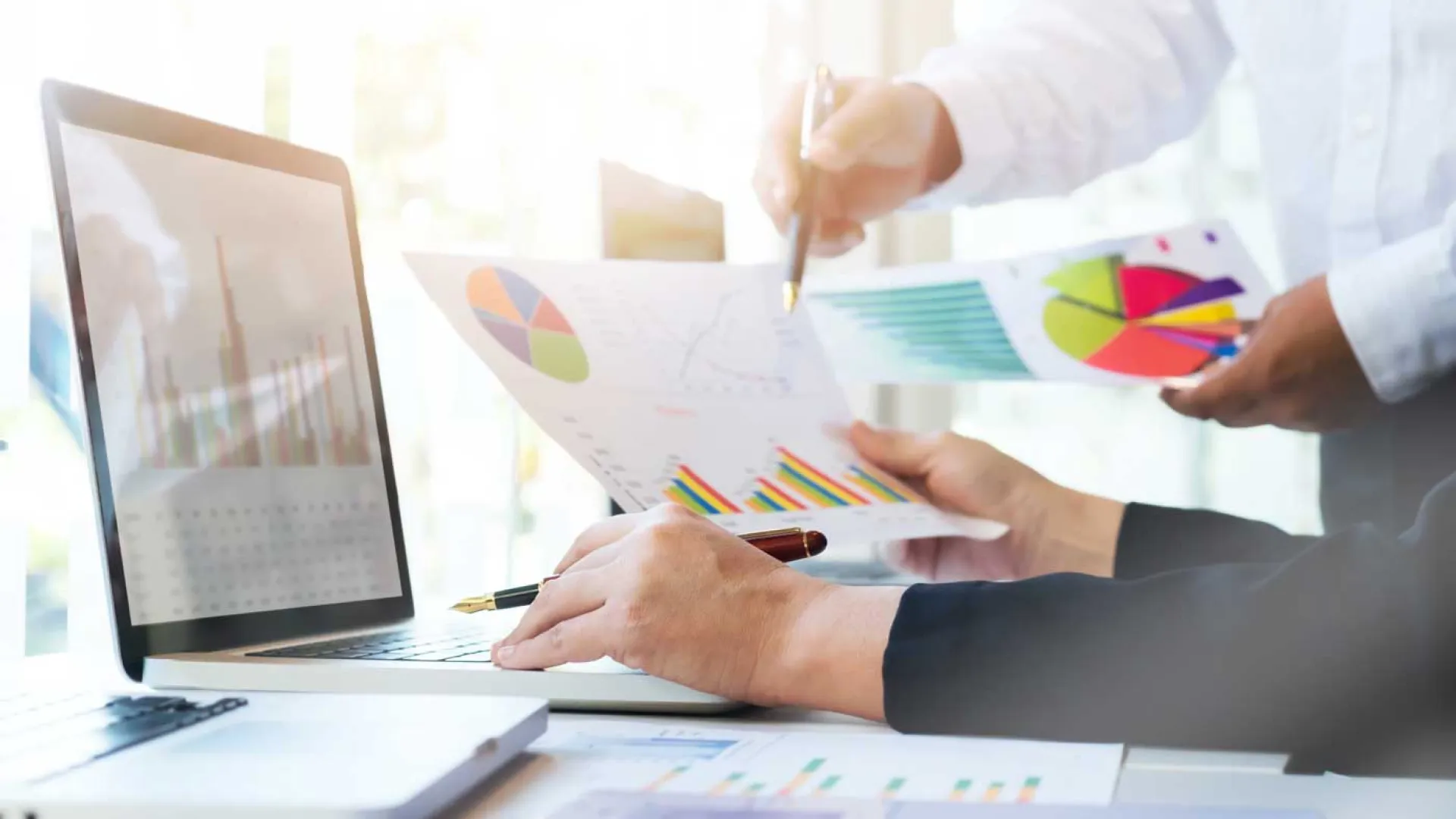Two professionals analyzing colorful charts and graphs on paper and a laptop during a business meeting.