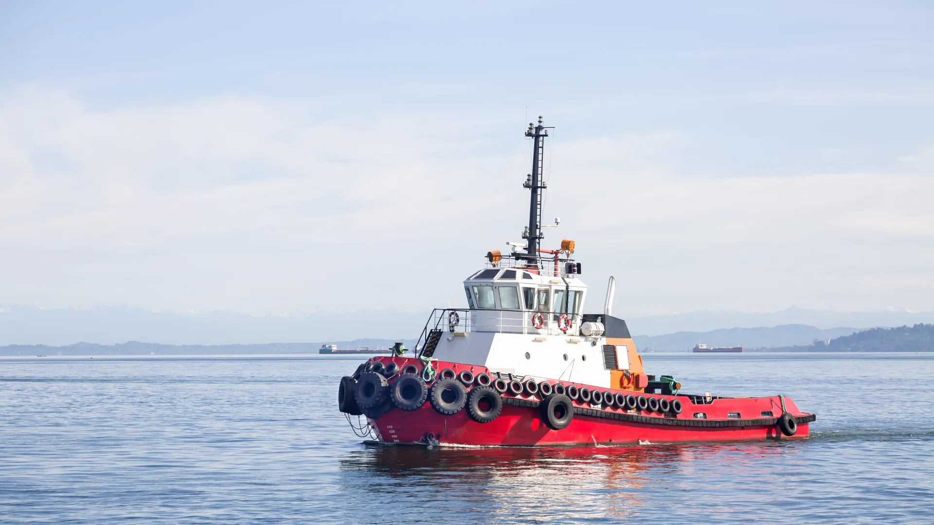 Red and white tugboat sailing on calm water with mountains and clear sky in the background.
