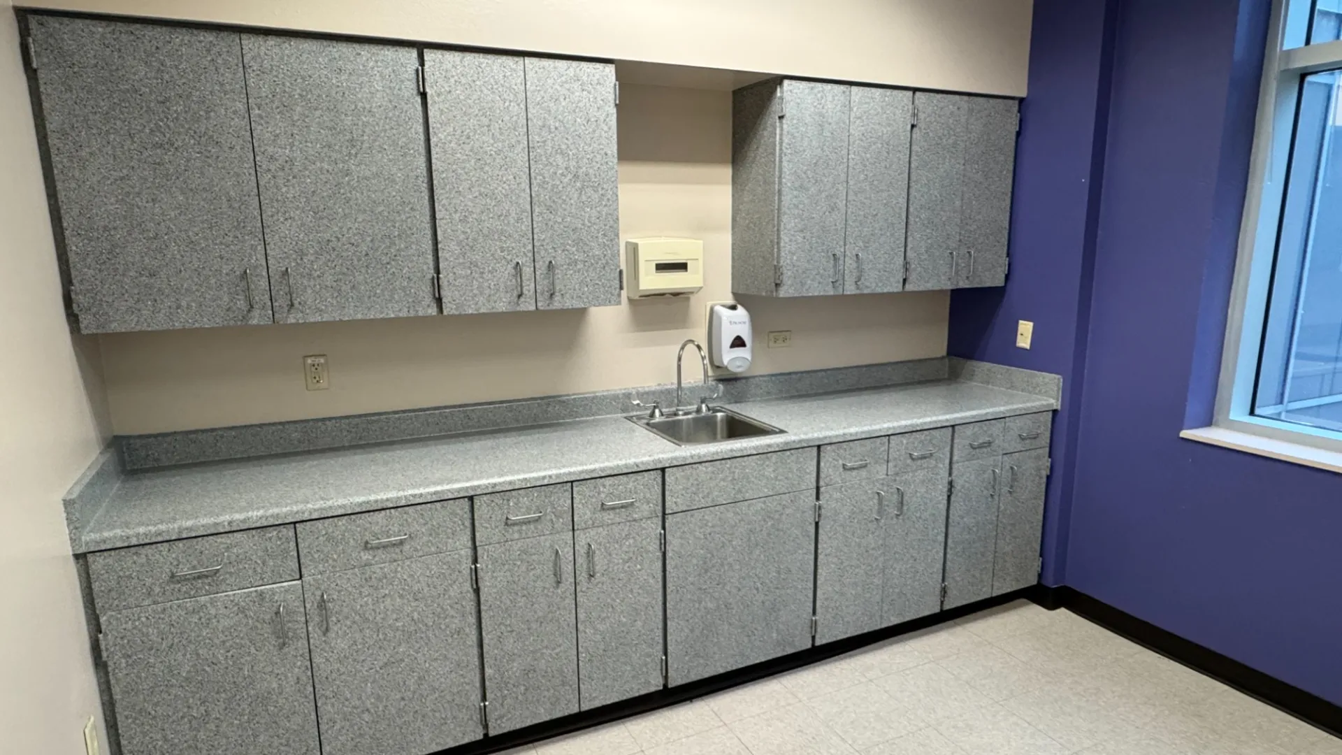 Empty room with gray cabinets, countertop, sink, and purple accented wall next to large window.