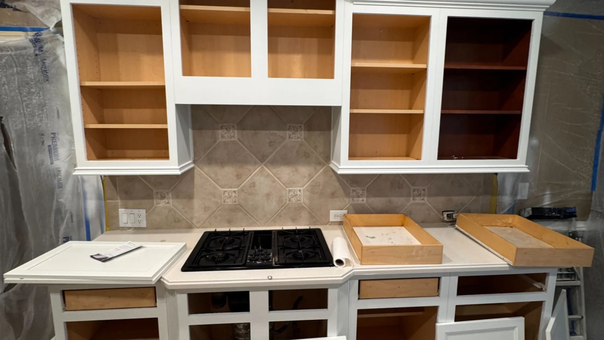 Unfinished white kitchen cabinets and gas stove installed during kitchen renovation with protective plastic sheeting.