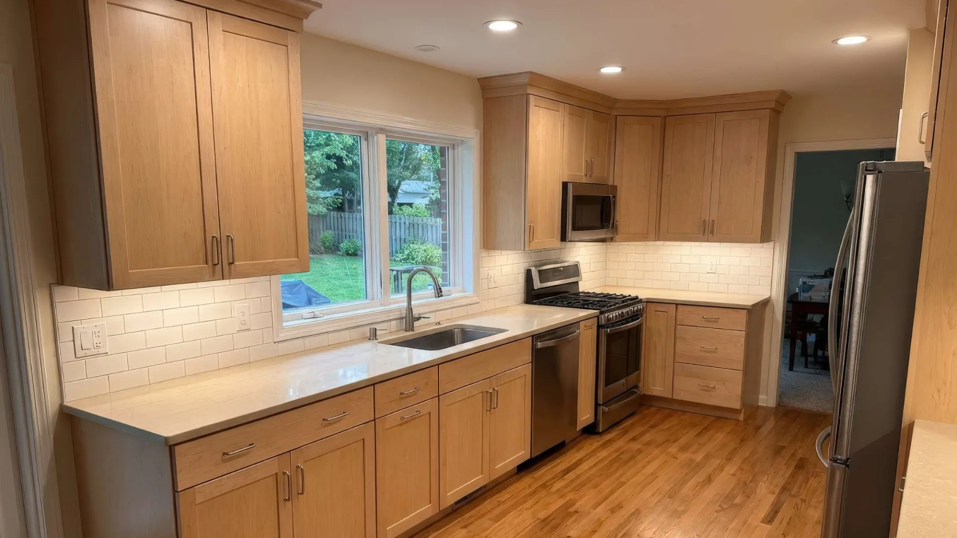 Modern kitchen with wooden cabinets, stainless steel appliances, hardwood flooring, and a large window over the sink.