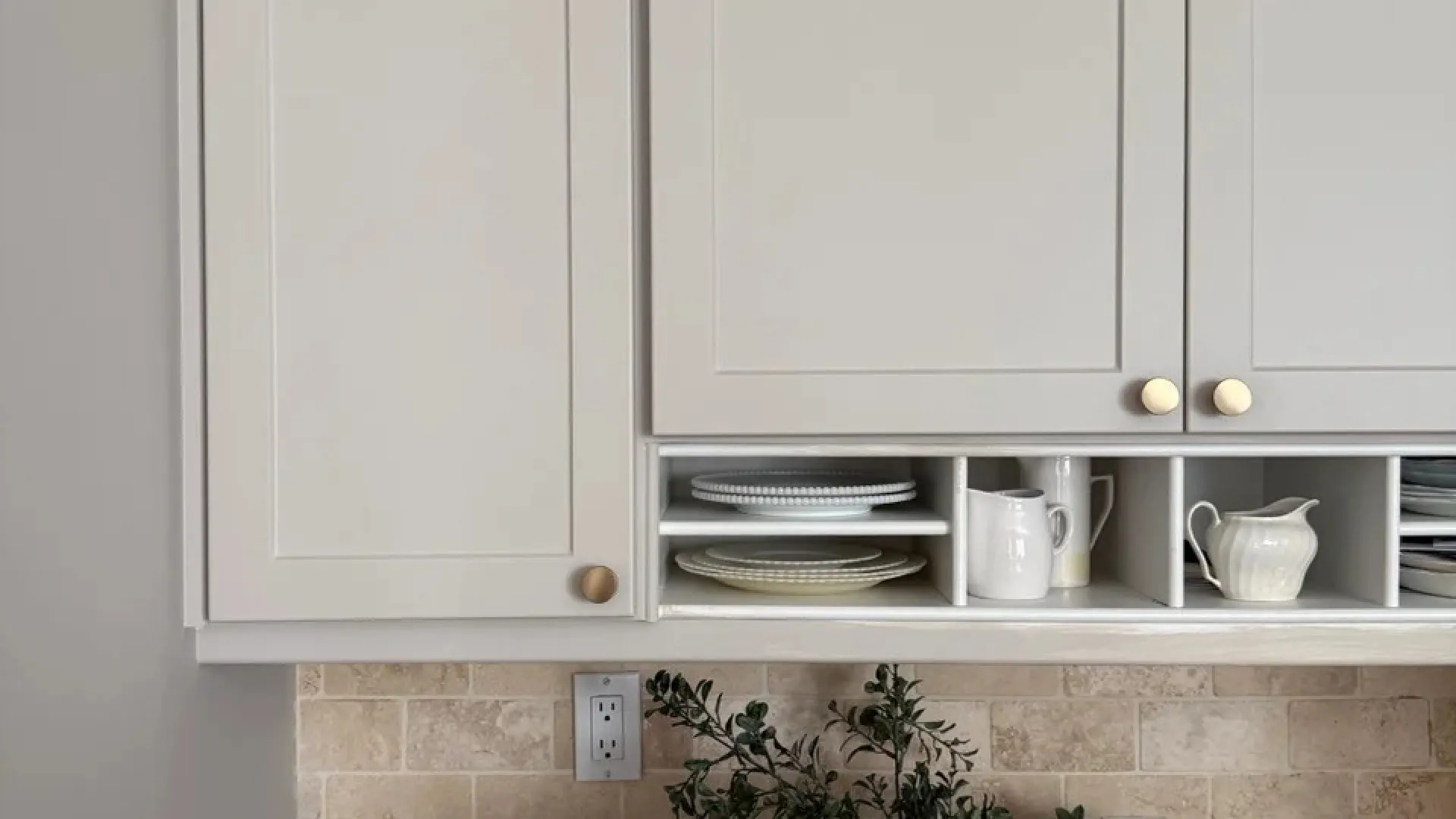 Neutral kitchen cabinets with open shelves holding dishes and a woven basket with greenery on a stone backsplash counter.