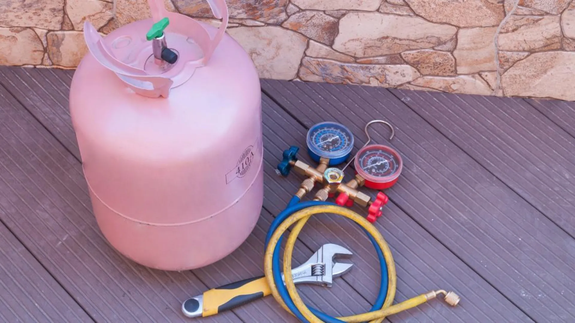 Pink refrigerant gas cylinder with manifold gauge set, hoses, and adjustable wrench on wooden floor near stone wall.