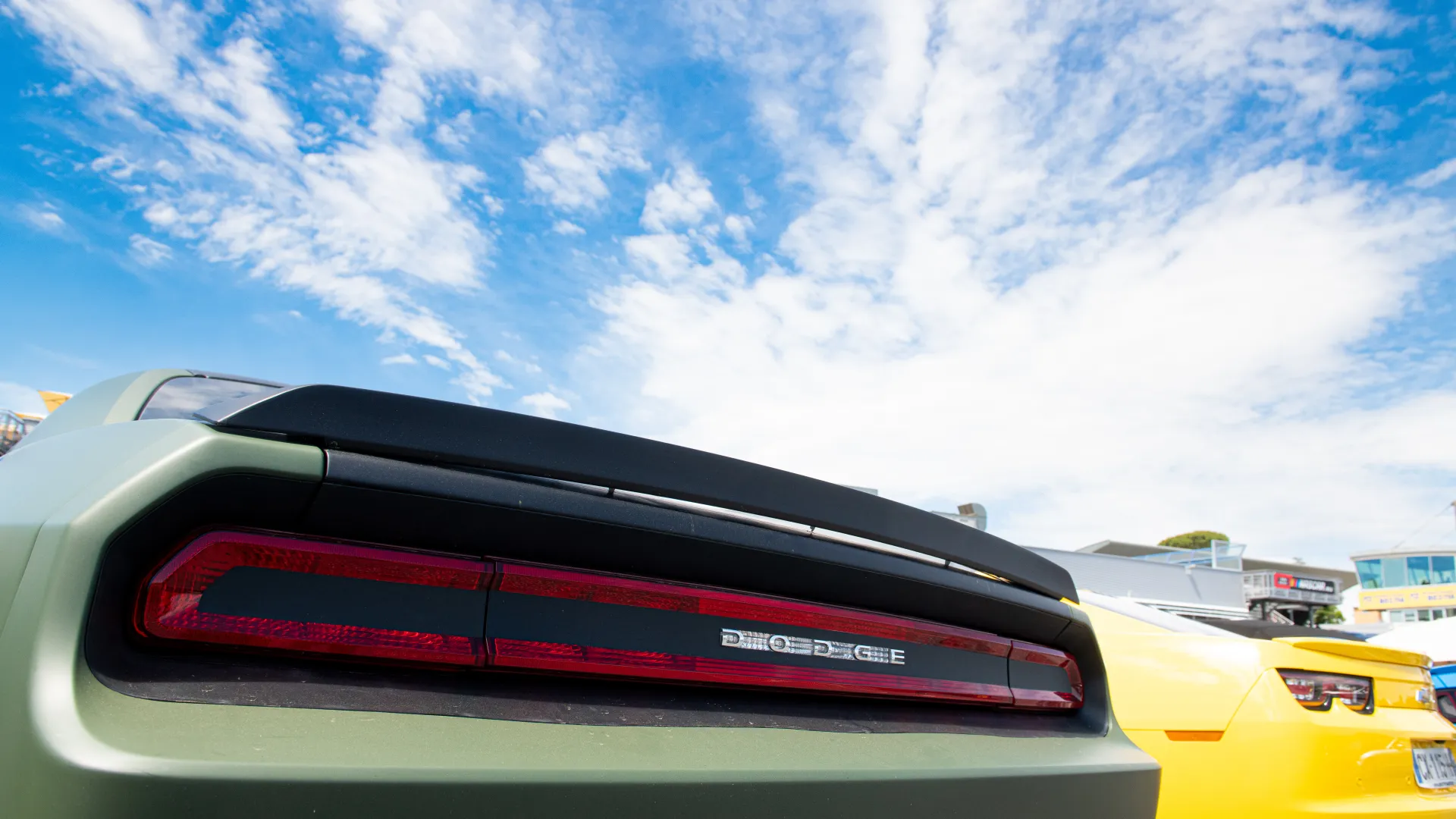 Close-up of a green Dodge car's rear with taillights under a blue sky with clouds, next to a yellow car.