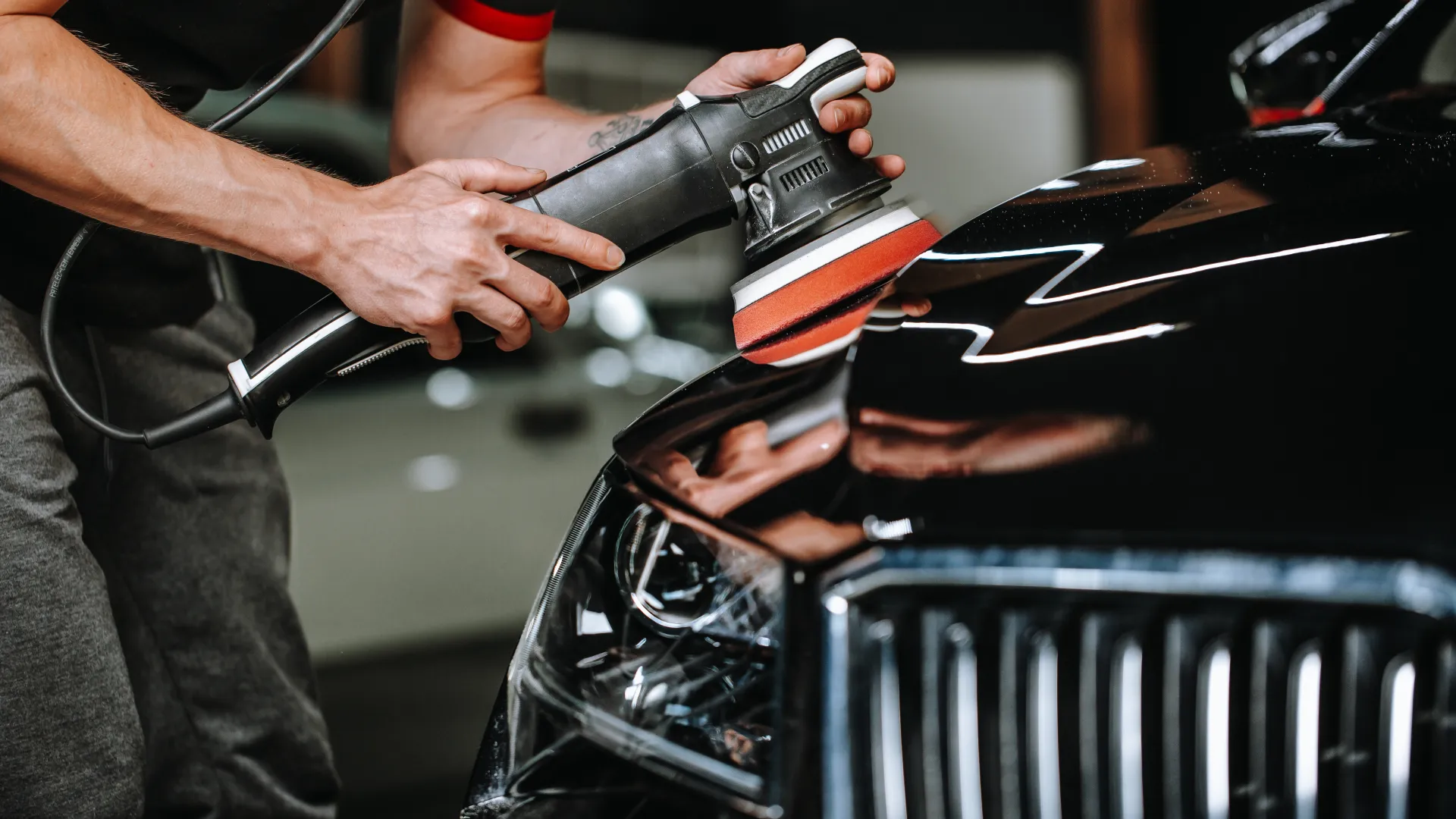 Person polishing black car hood using electric buffer in a garage or detailing shop setting.