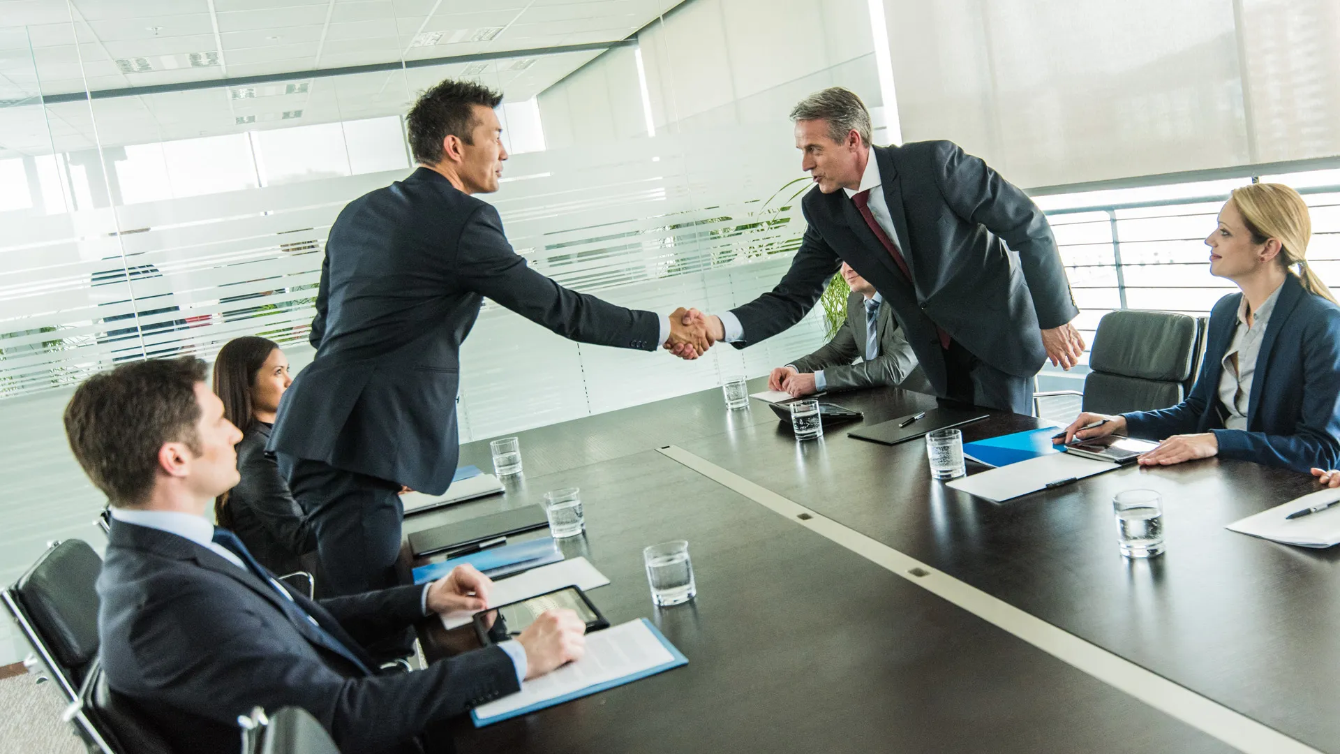 Two businessmen shaking hands across a conference table while colleagues observe in a modern office.